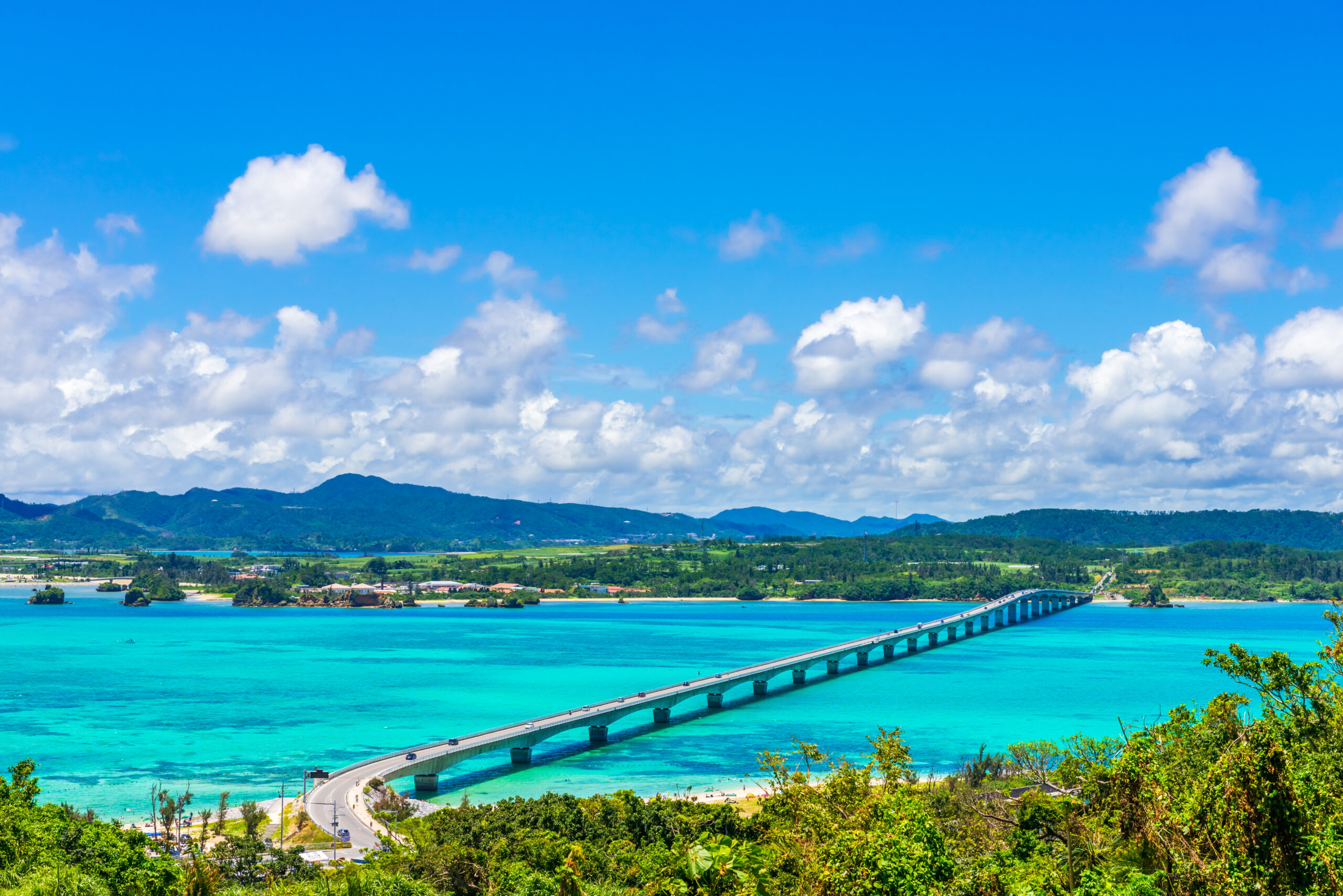 Bridge extends over bright turquoise water toward a distant shoreline with green hills and a blue sky filled with fluffy clouds, suggesting a coastal landscape.