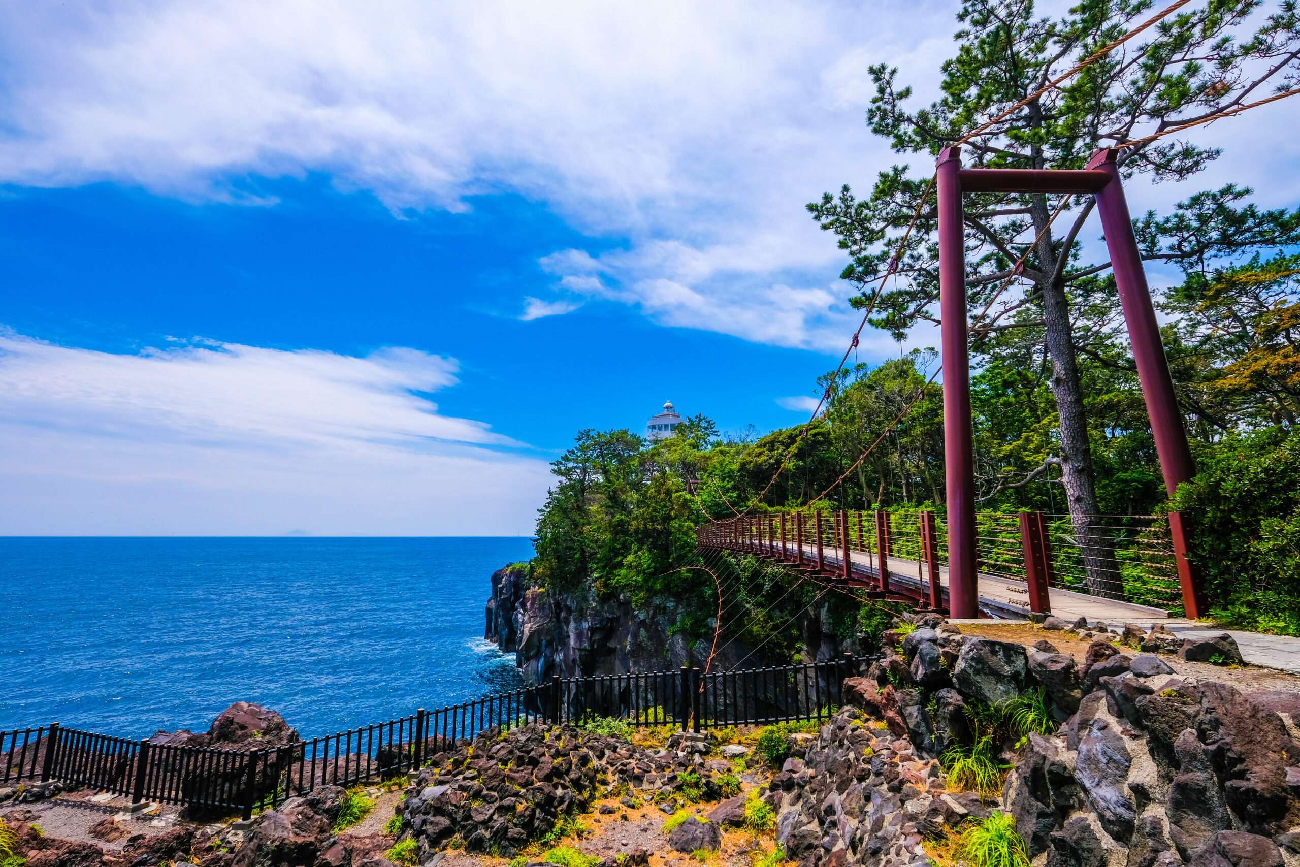 Coastal walkway along a rocky shore with red posts, rope railings, pine trees, and a blue sea under a bright sky.