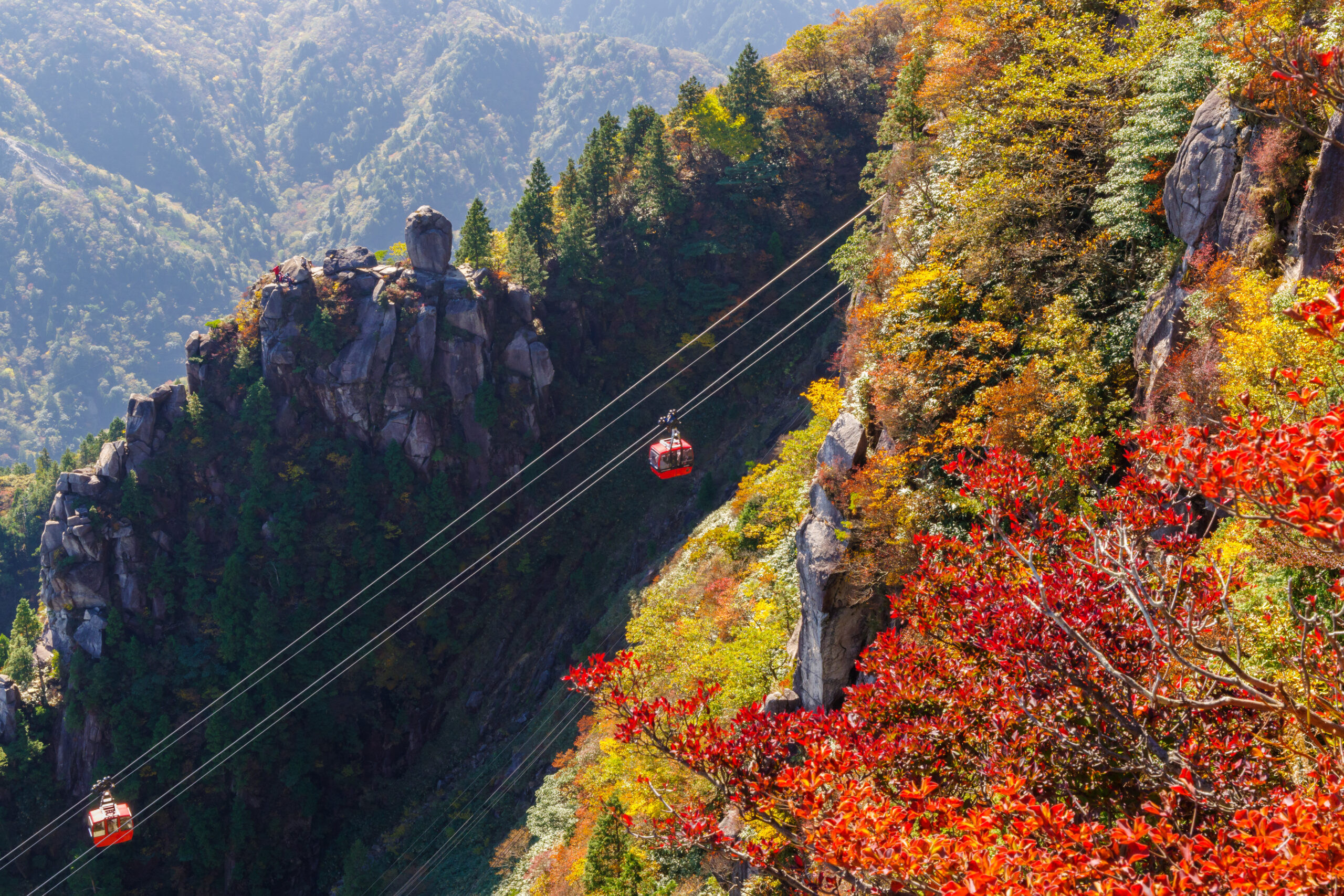 Red cable car glides along steel cables over a deep valley, surrounded by autumn-colored trees and rugged cliffs.