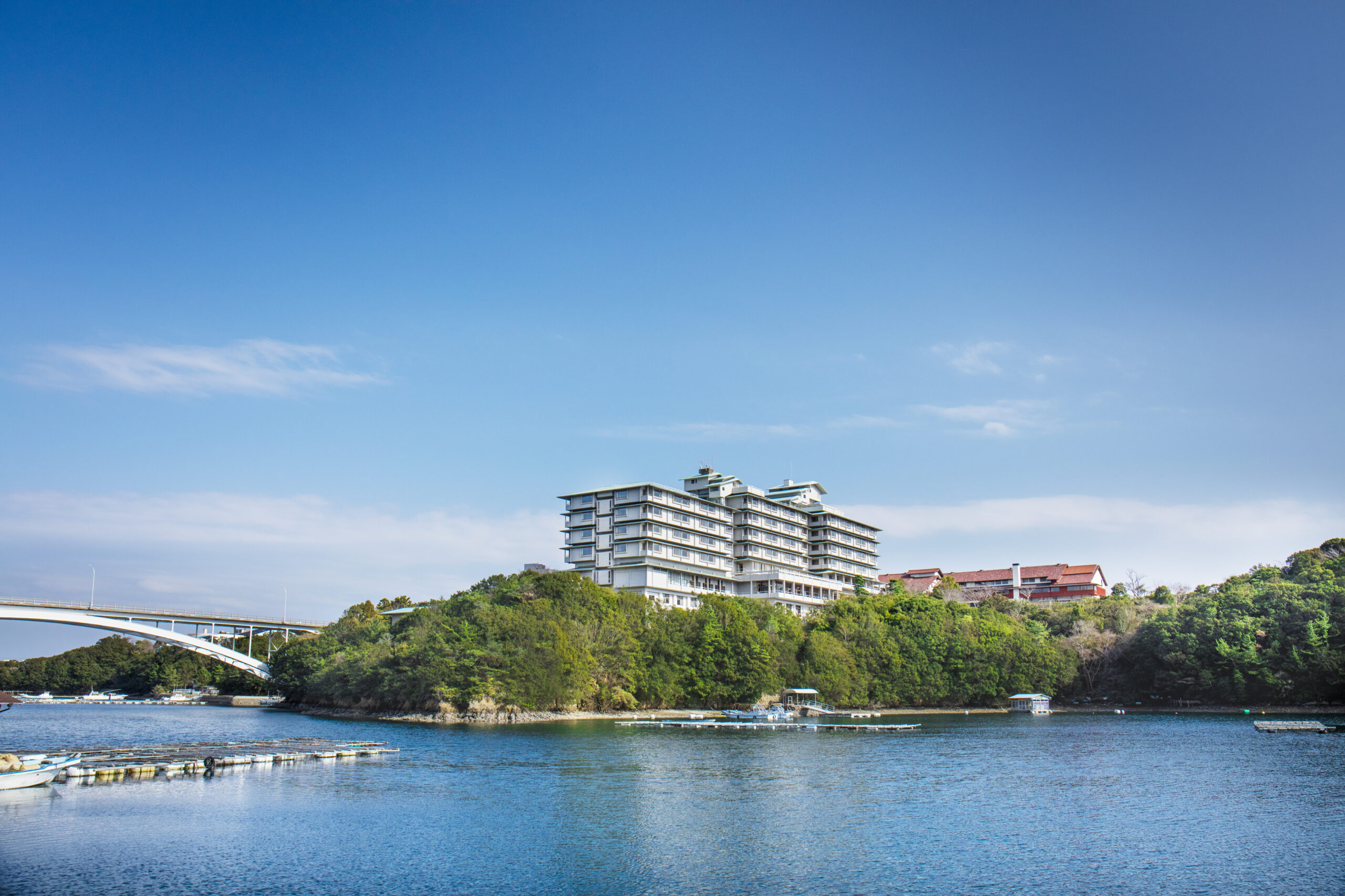 Modern white apartment building perched on a hill above a blue river, with a curved bridge on the left and green trees surrounding the shore.