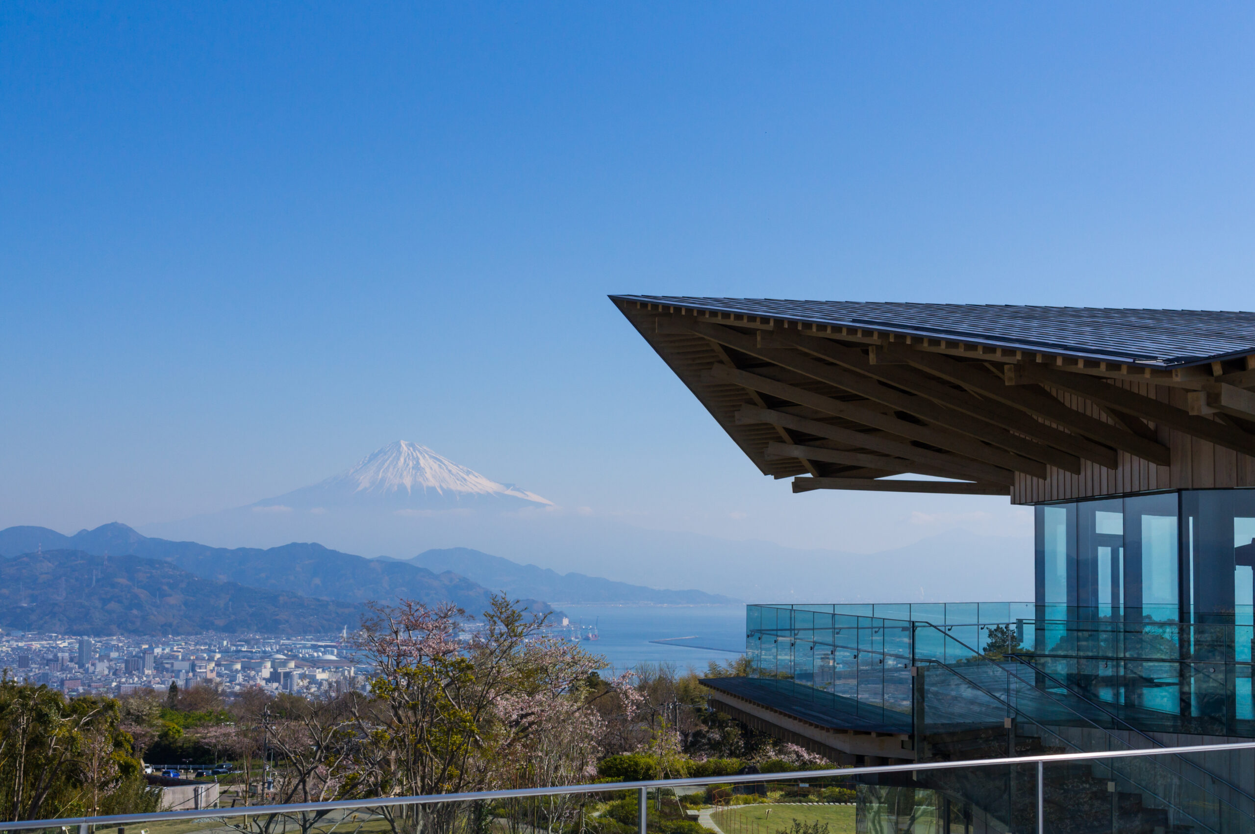 Modern wooden pavilion extends over glass railing with a city below and a snow-capped mountain in the distance under a clear blue sky