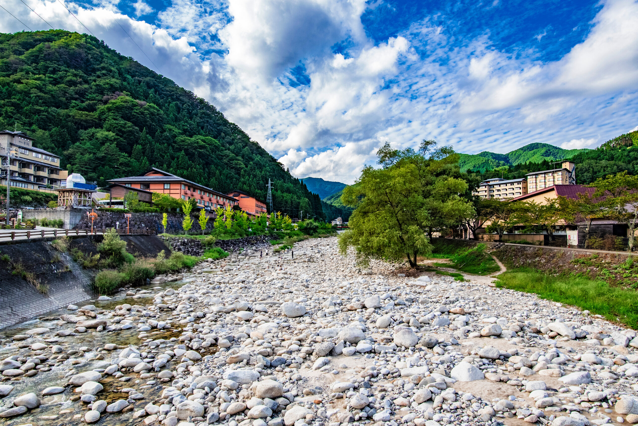 Rocky riverbed with a shallow stream and green, forested mountains and buildings along the shore under a blue, cloudy sky?