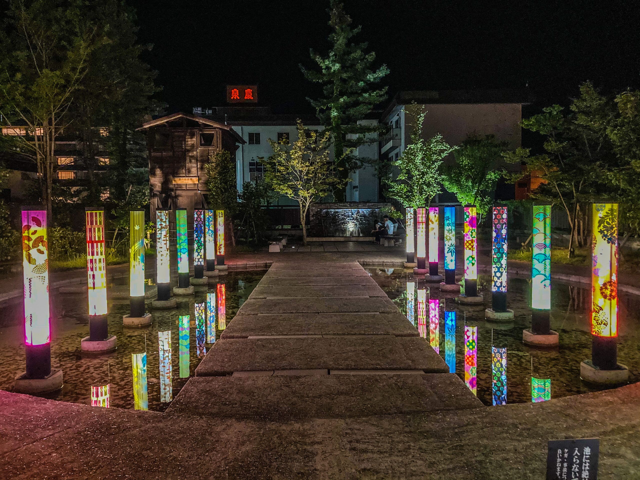 Night scene of a walkway over water bordered by tall illuminated colorful vertical banners with a dark sky above.
