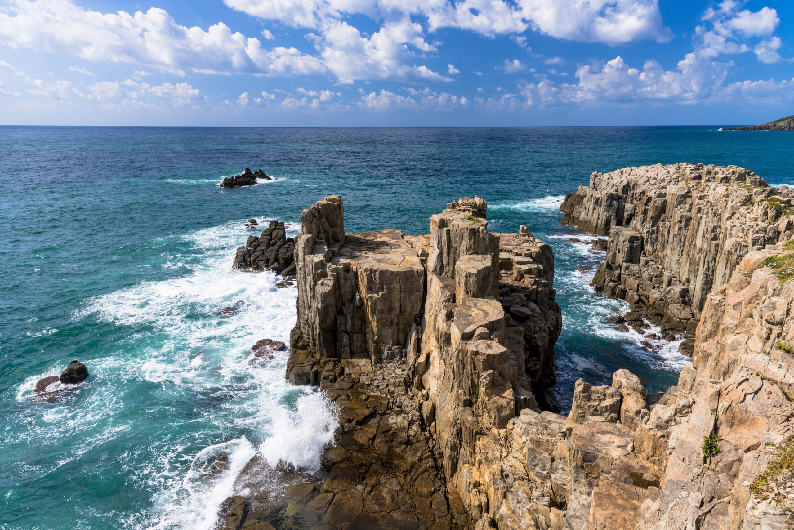 Jagged rocky shoreline with turquoise water crashing against cliffs under a blue sky with white clouds, coastal scene.