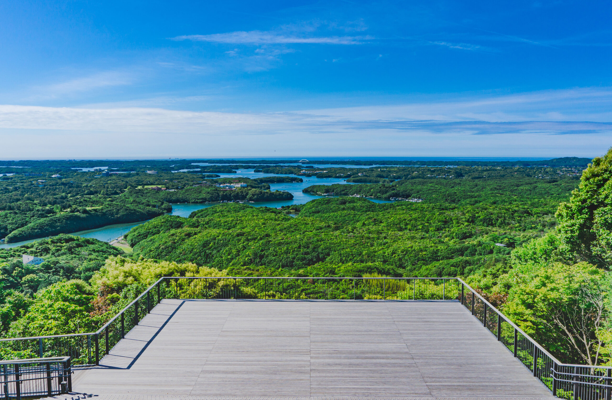 Wooden observation deck with metal railing overlooking dense green hills, water beyond, and a clear blue sky.