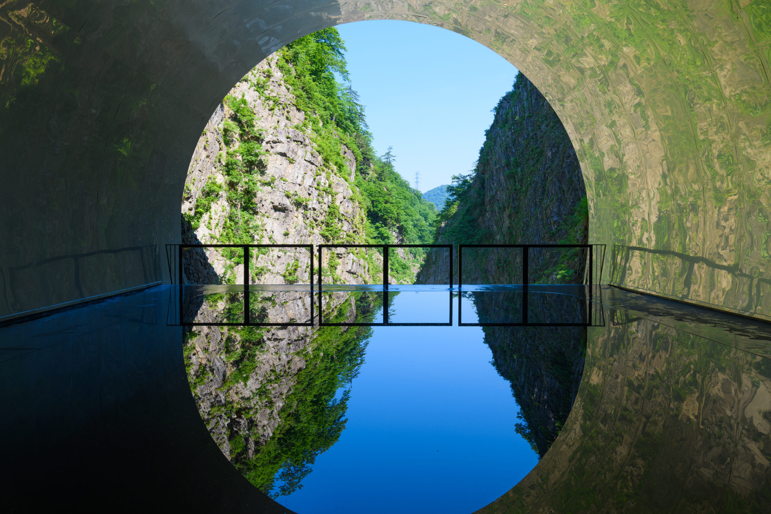 Circular tunnel opening revealing a rocky, green canyon and blue sky; railing along a reflective pool inside the tunnel.