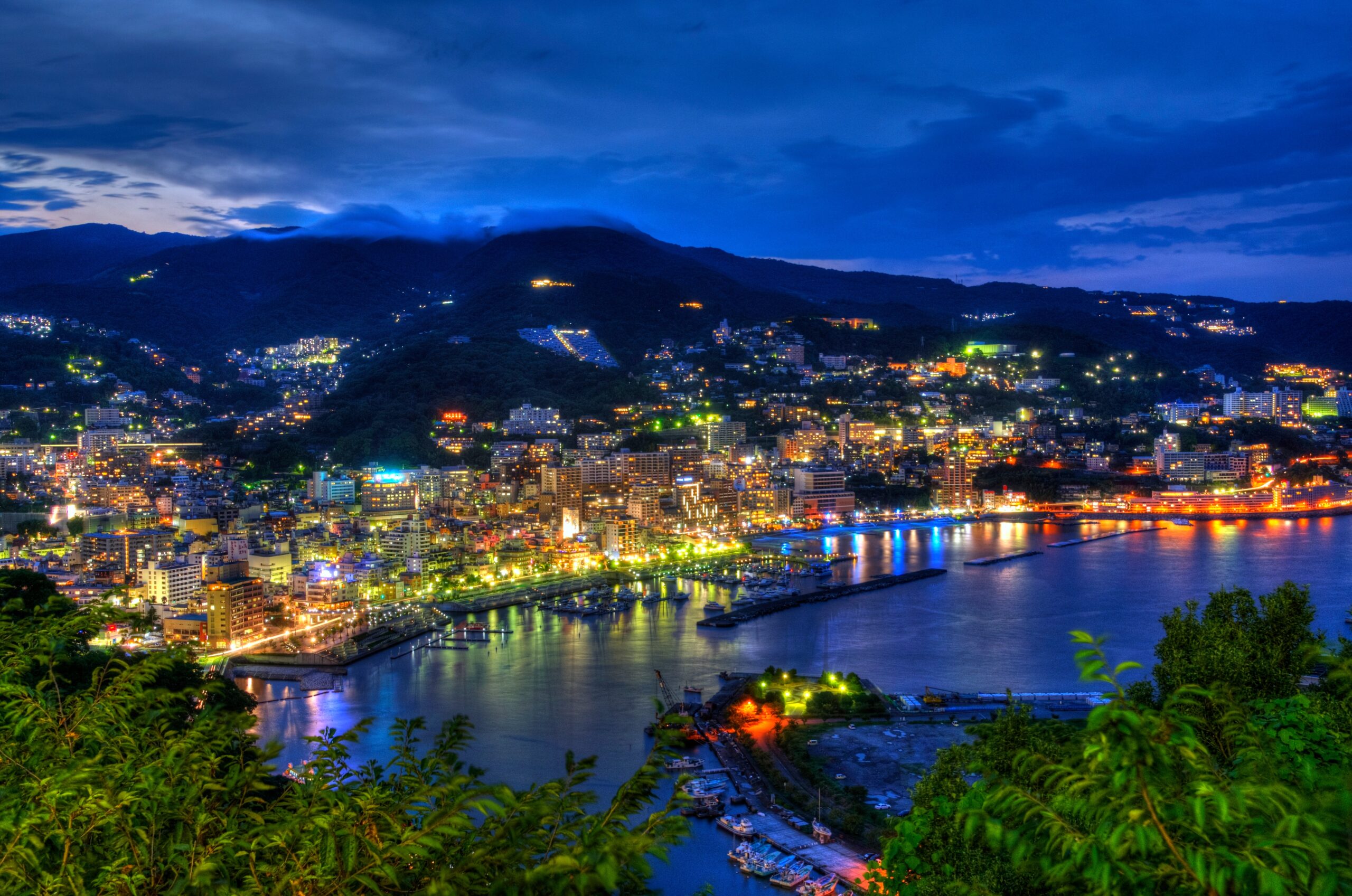 Nighttime cityscape along a bay with illuminated buildings, hills, and a calm harbor under a deep blue sky