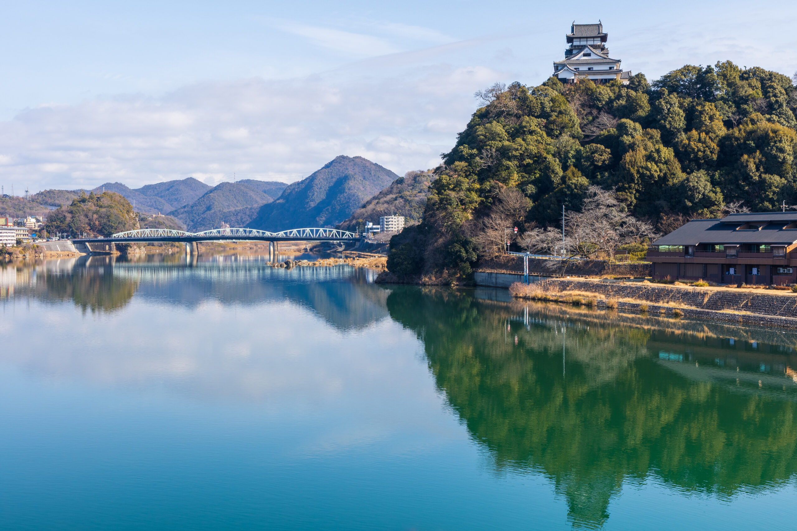Temple perched on a forested hill above a calm river with a white arched bridge in the distance.