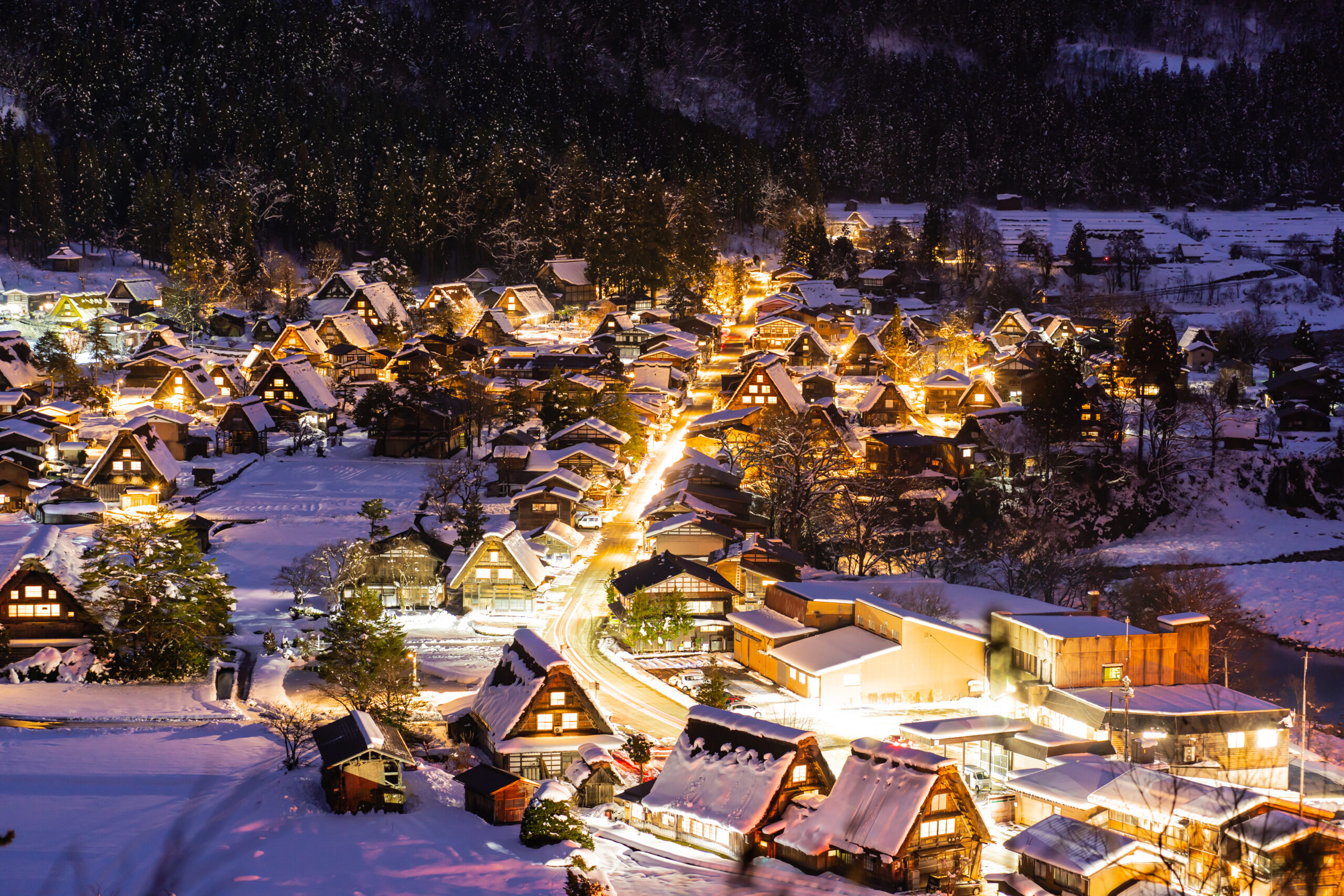 Aerial view of a snow-covered village at night, warm lights glowing from chalet-style houses along winding streets.