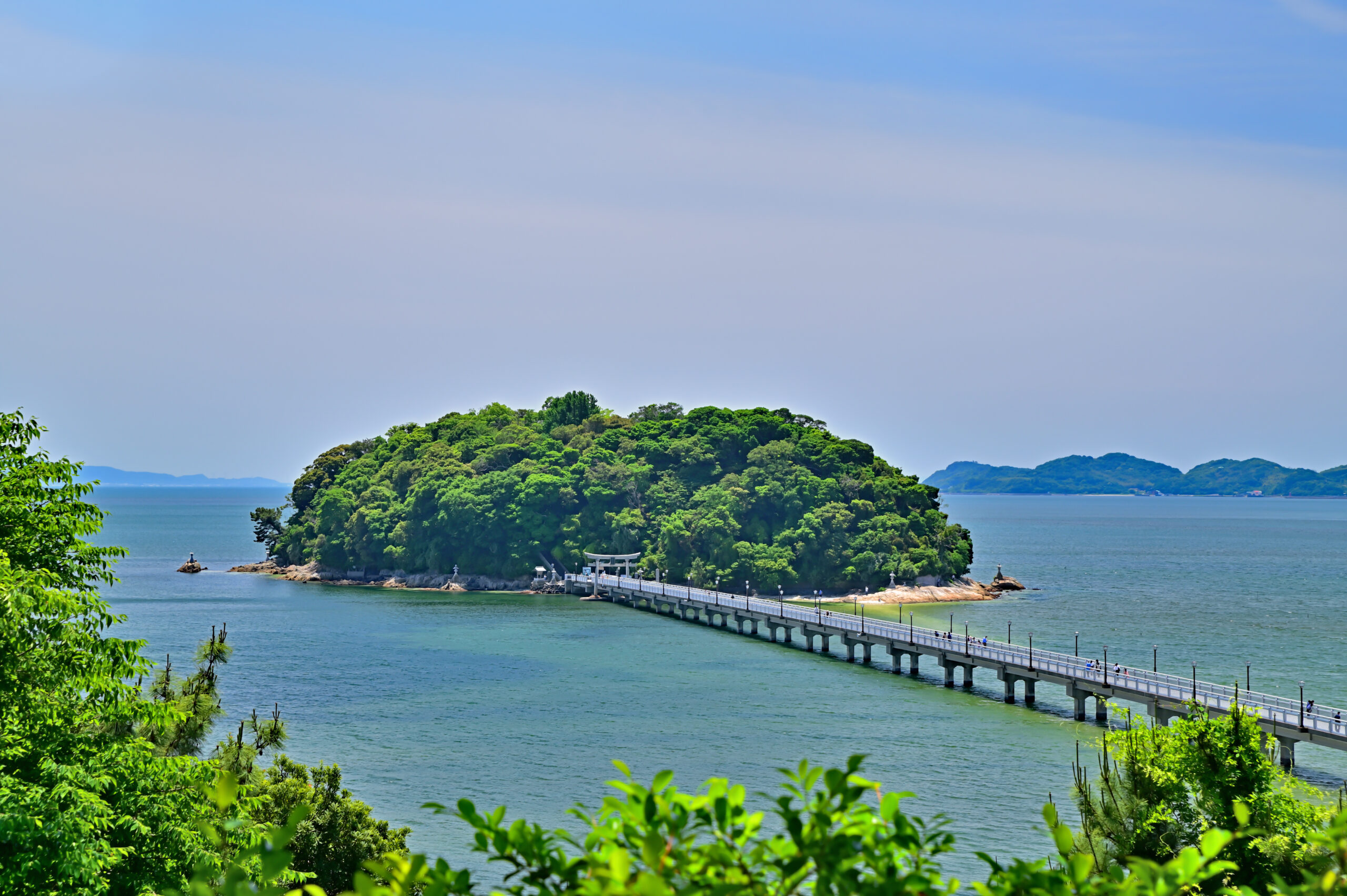 Island densely covered with trees connected to the mainland by a long bridge over calm blue water, with people walking along it.