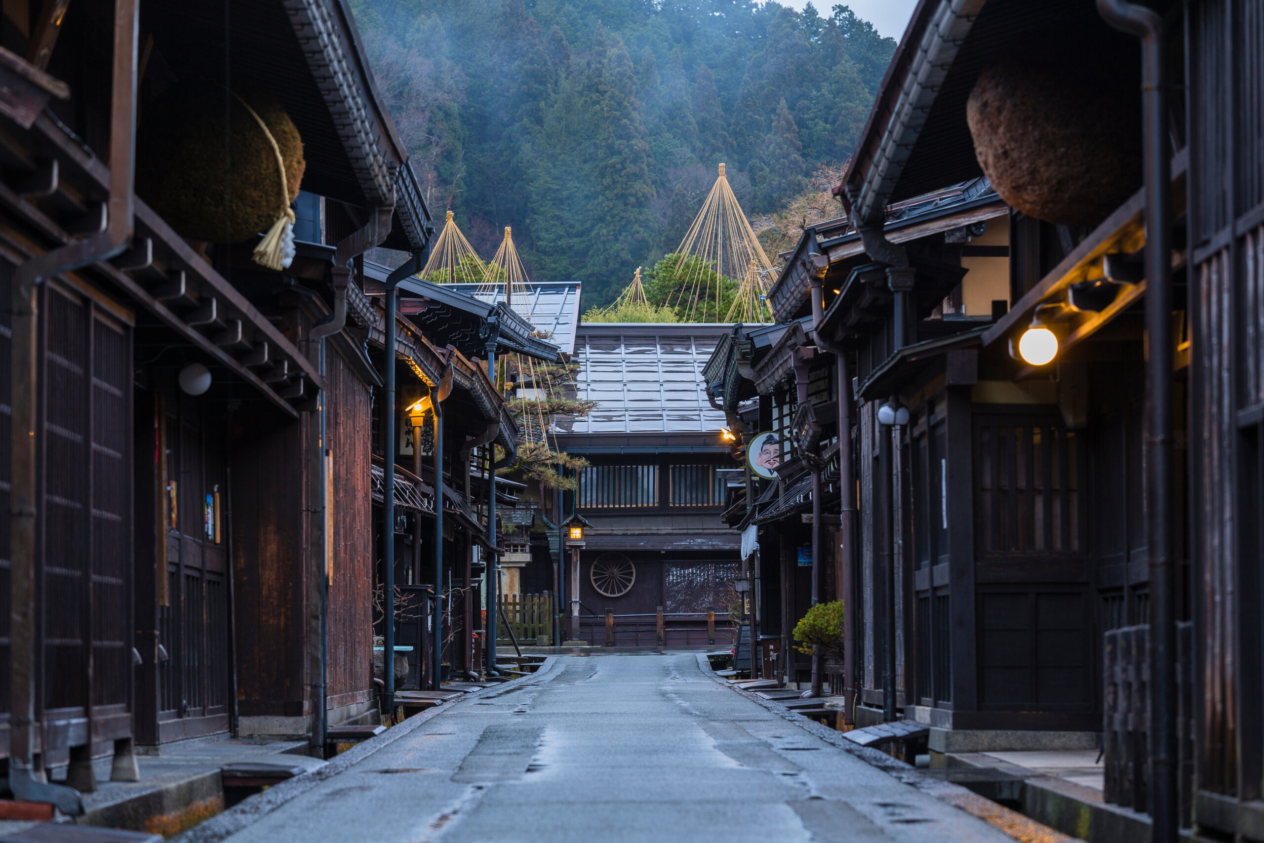 Narrow street in a traditional Japanese town with dark wooden storefronts and glowing lanterns, mountain backdrop in the distance.