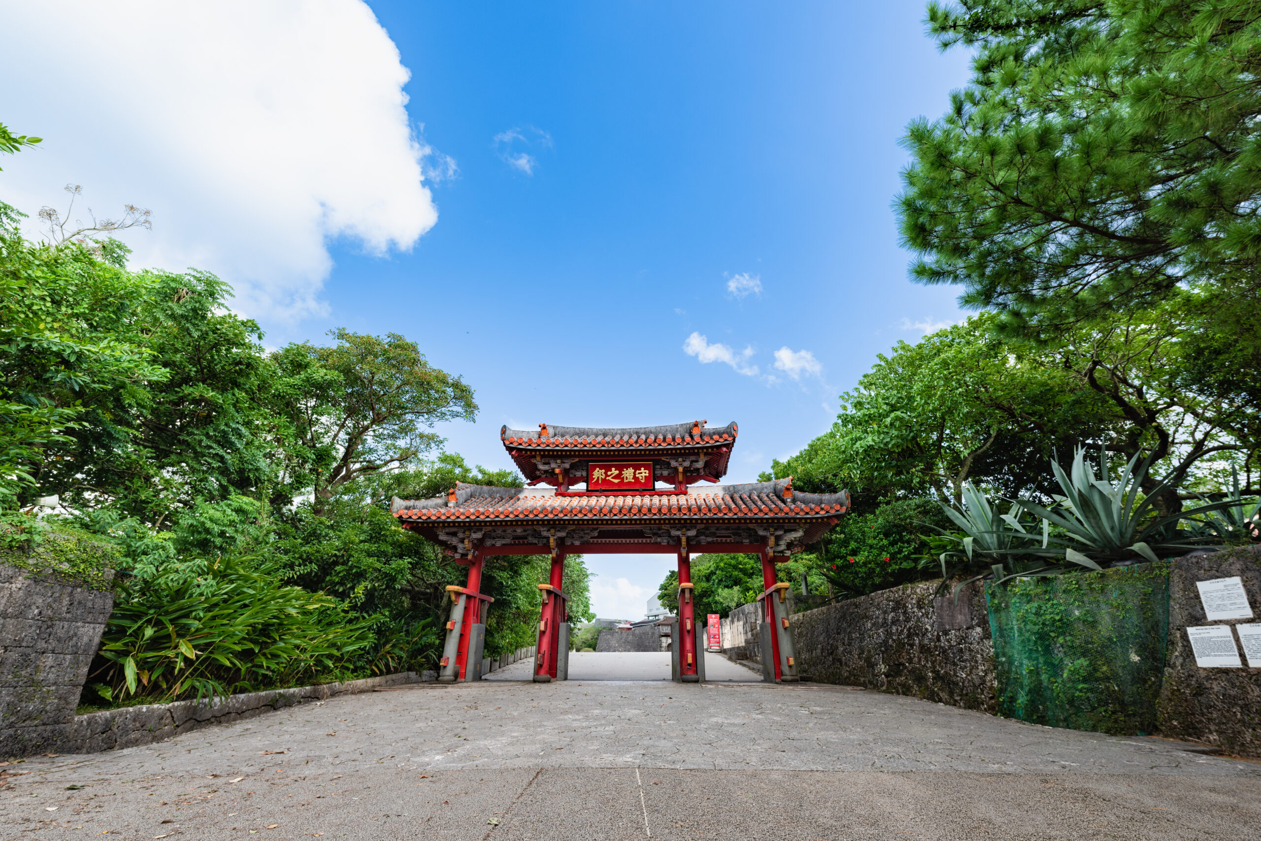 Red Chinese gate with ornate tiled roof framed by lush green trees and a bright blue sky.