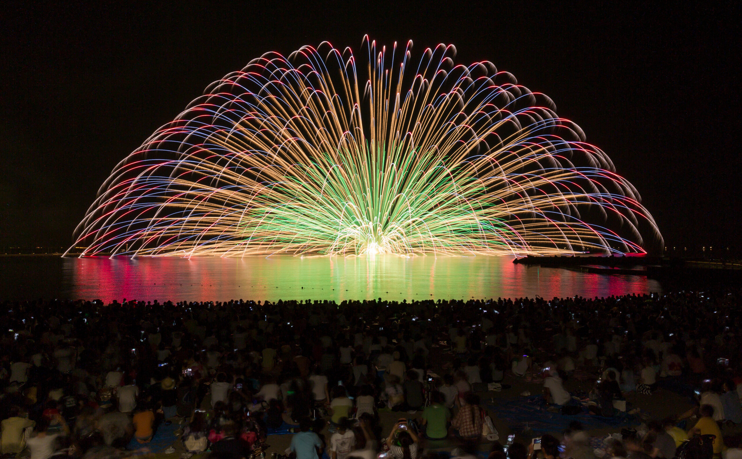 Nighttime fireworks over a lake, colors reflecting on the water, with a large crowd watching from the shore.