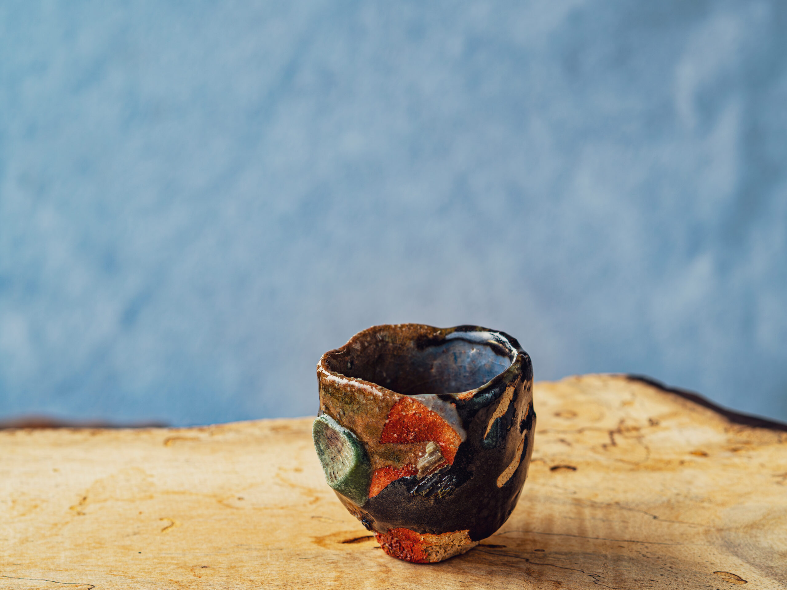 Close-up of a small, irregular ceramic mug with red, green glaze on a weathered wood surface under a blue sky.