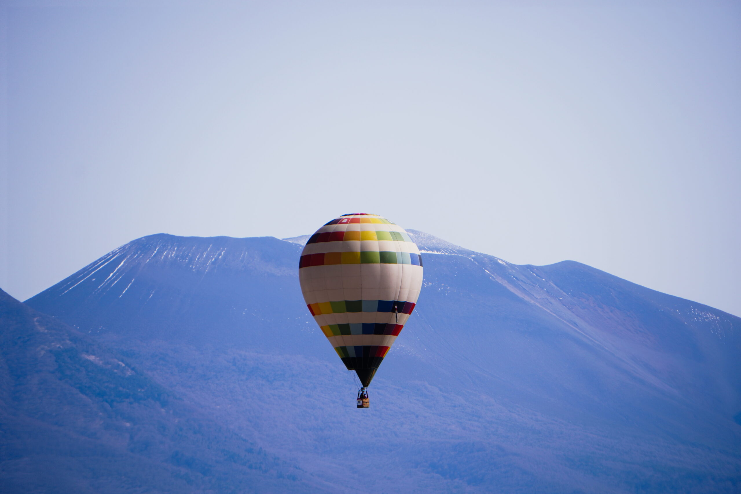 Colorful hot air balloon floating over snow-dusted mountains under a clear blue sky with a distant valley below