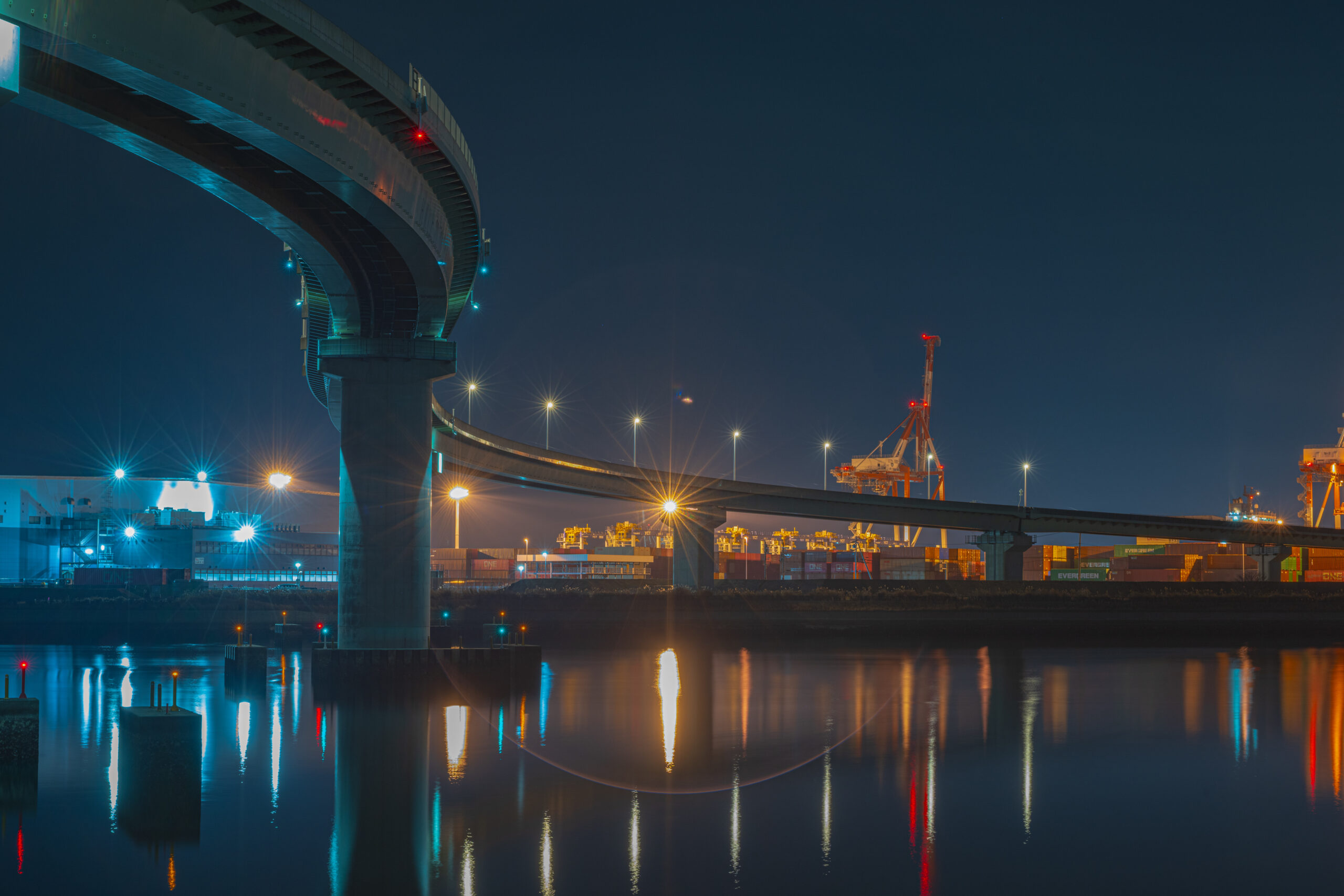 Night view of a curved elevated highway over water with bright streetlights and blue-green illumination, port cranes visible in the distance reflecting in the water