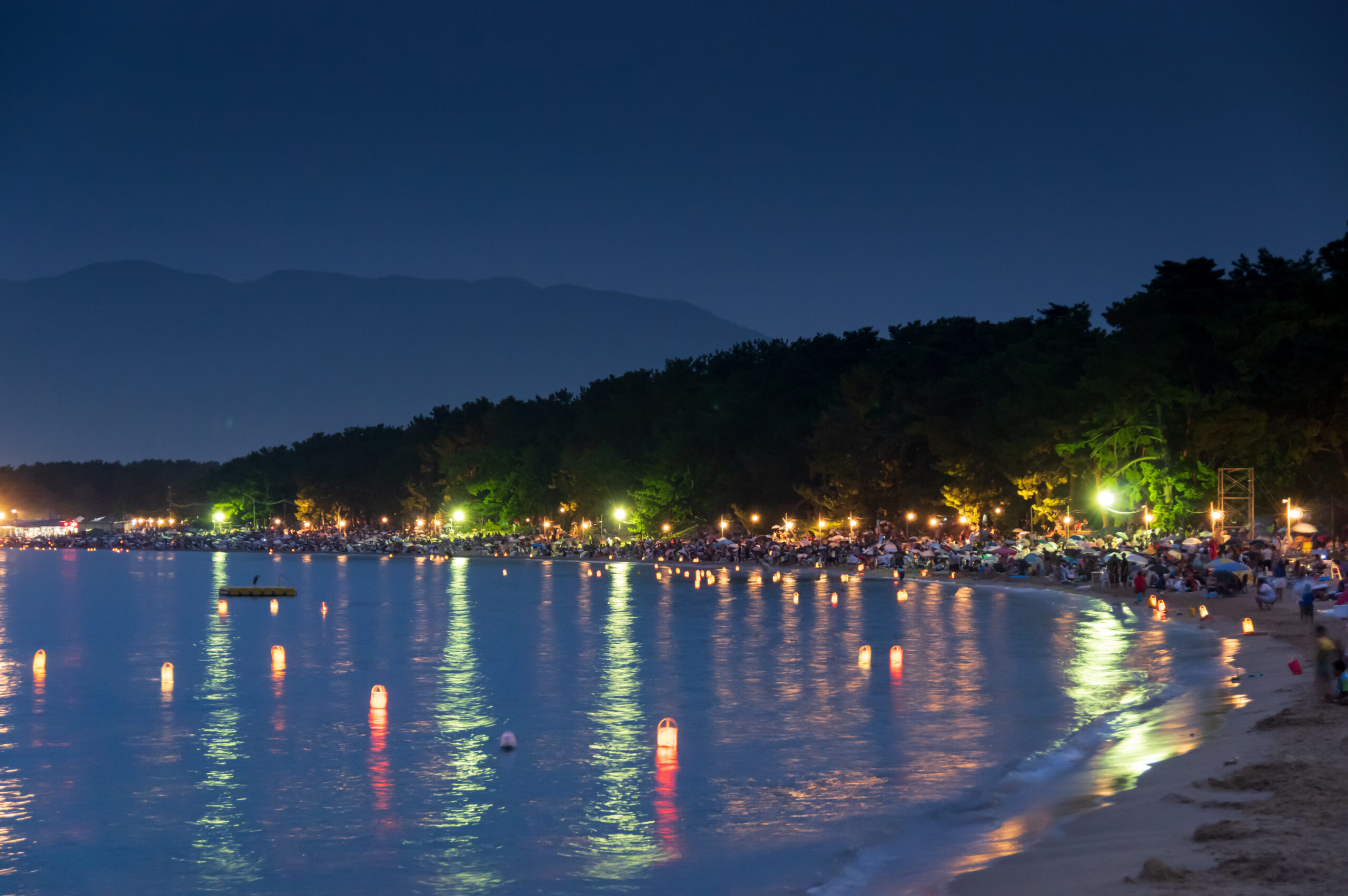 Crowded beach at night with numerous glowing lanterns floating on calm water and distant hills in the background.