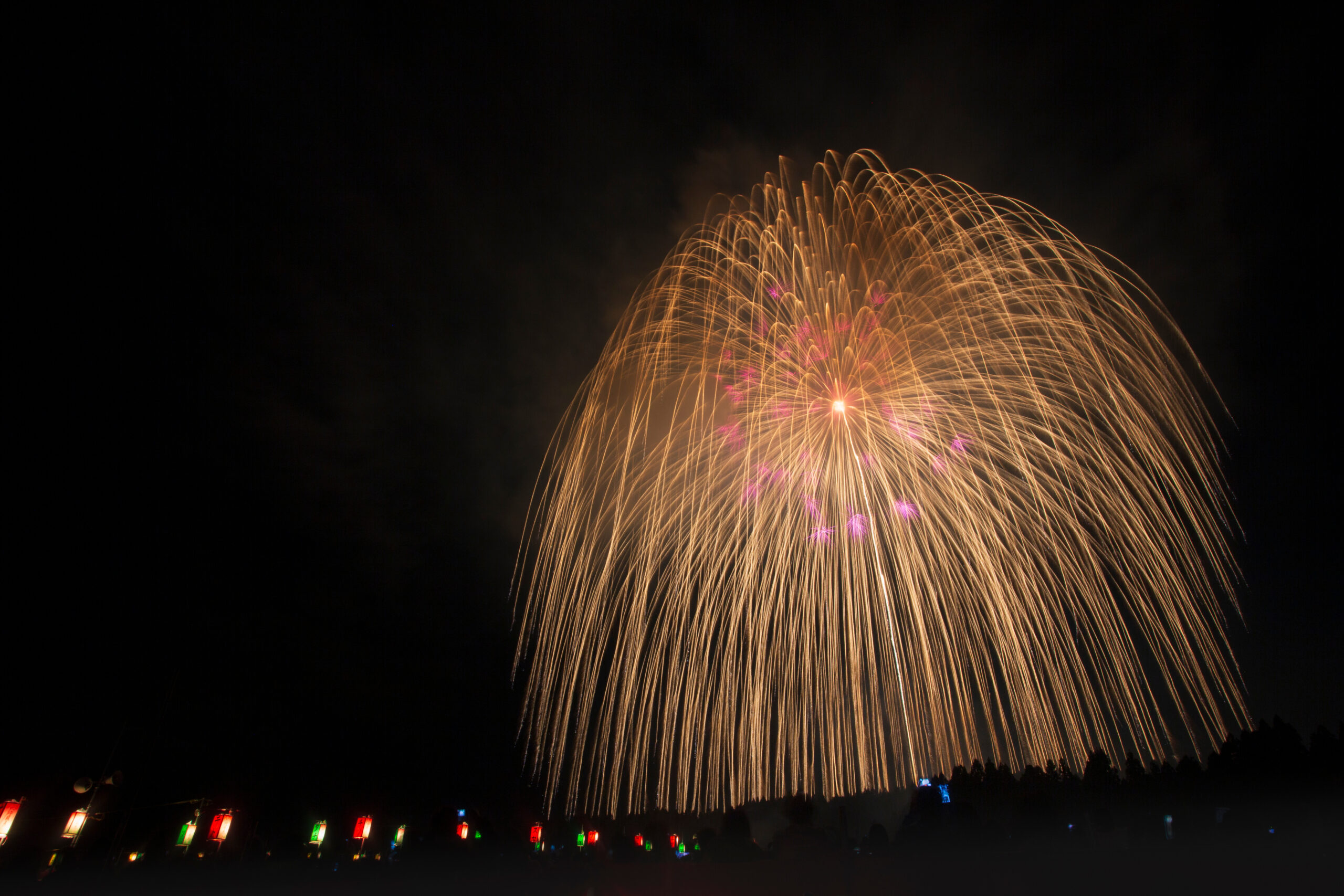 Golden fireworks burst in an arc across the night sky over a festival crowd, with colorful lanterns glowing along the ground below.