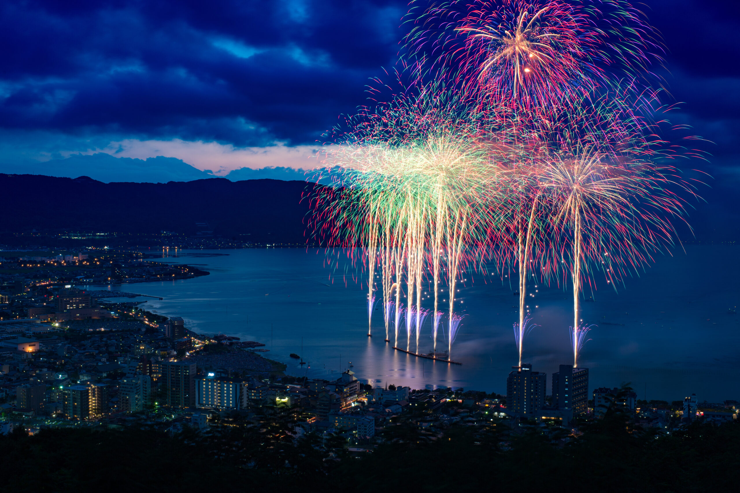 Colorful fireworks lighting up the night sky over a coastal city with hills in the background and illuminated buildings along the water