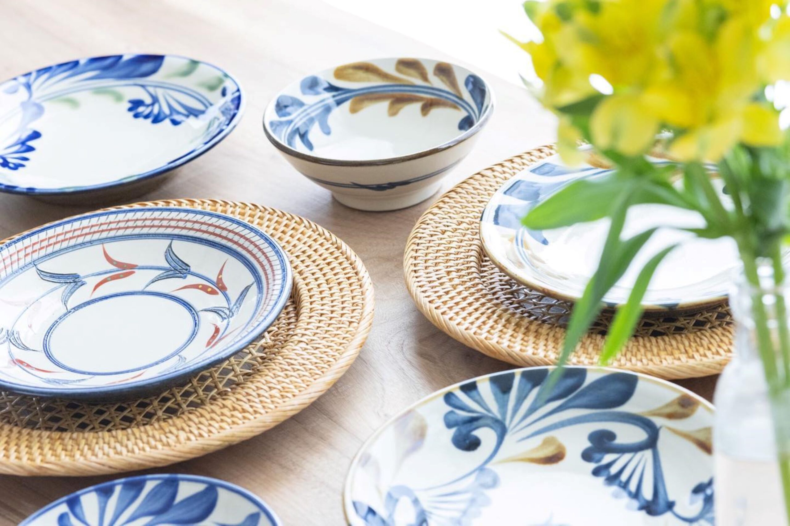 Blue and white ceramic plates with floral patterns on woven placemats, arranged for a table setting with greenery in the background.