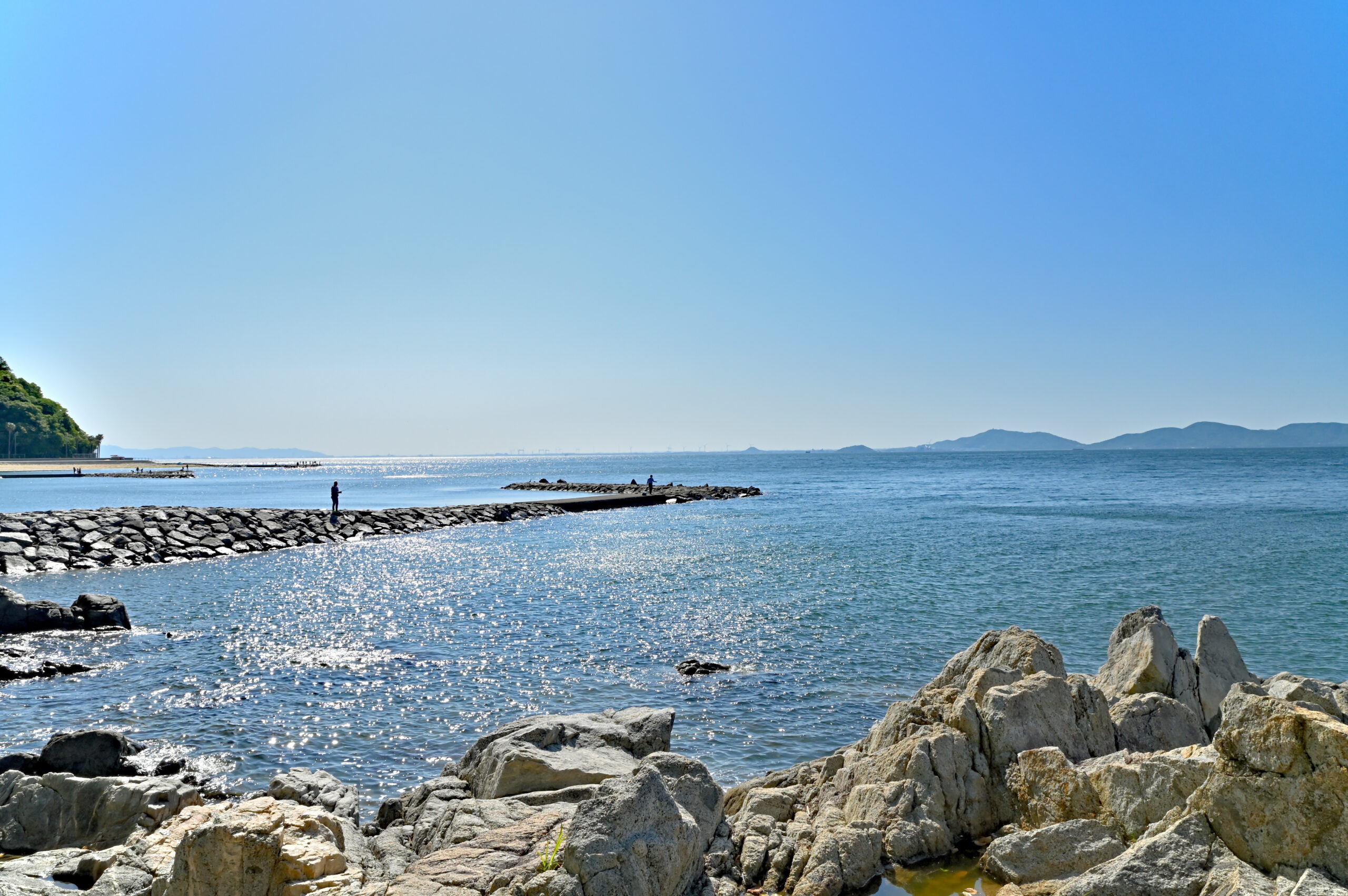 Calm blue sea with a rocky foreground and a stone jetty where a few people are fishing under a clear sky.