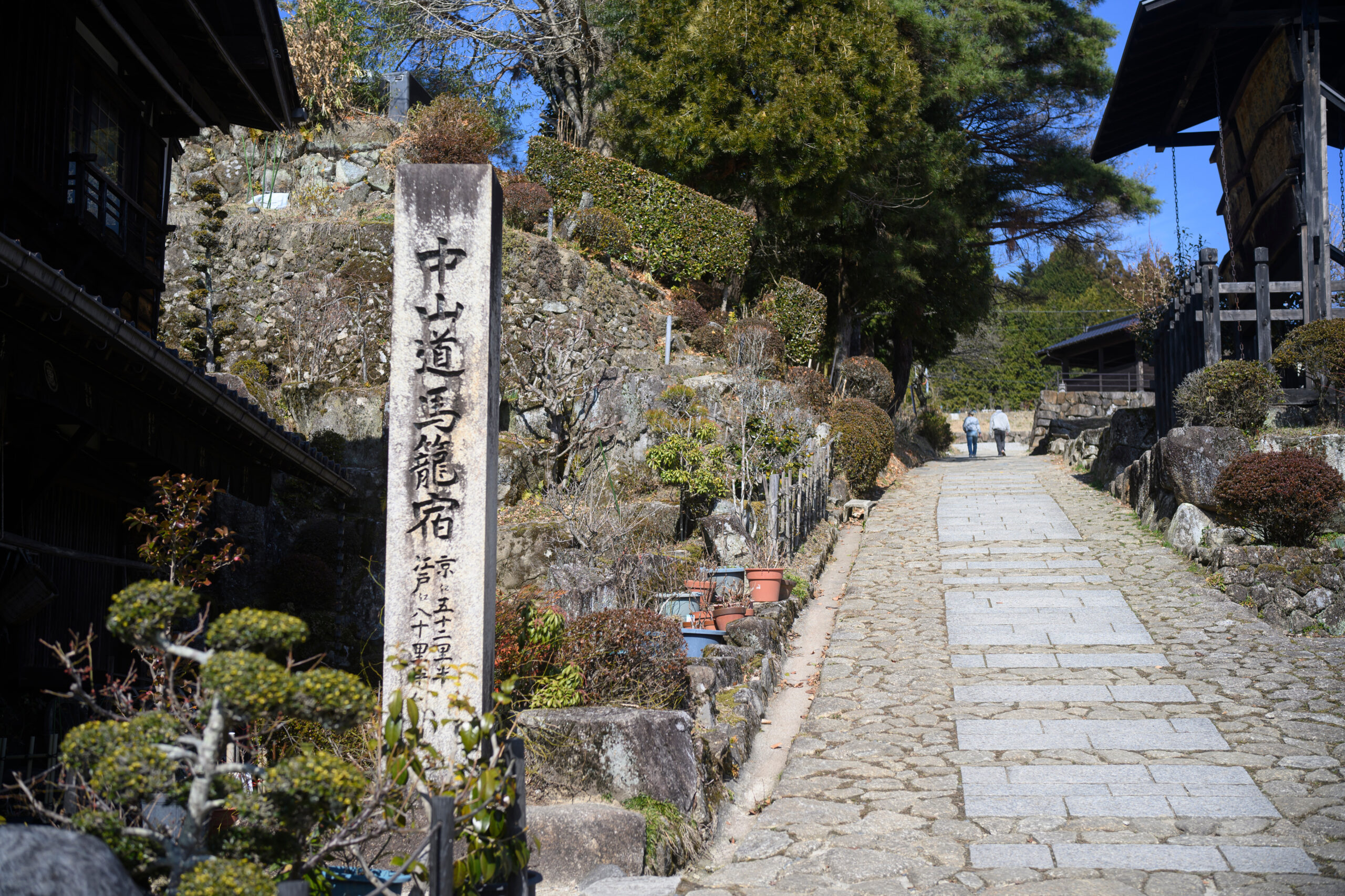 Stone-paved street in a Japanese temple or village, with a tall inscribed stone marker on the left, and traditional wooden buildings along a stone wall and greenery under a clear blue sky. Two people walk away in the distance.
