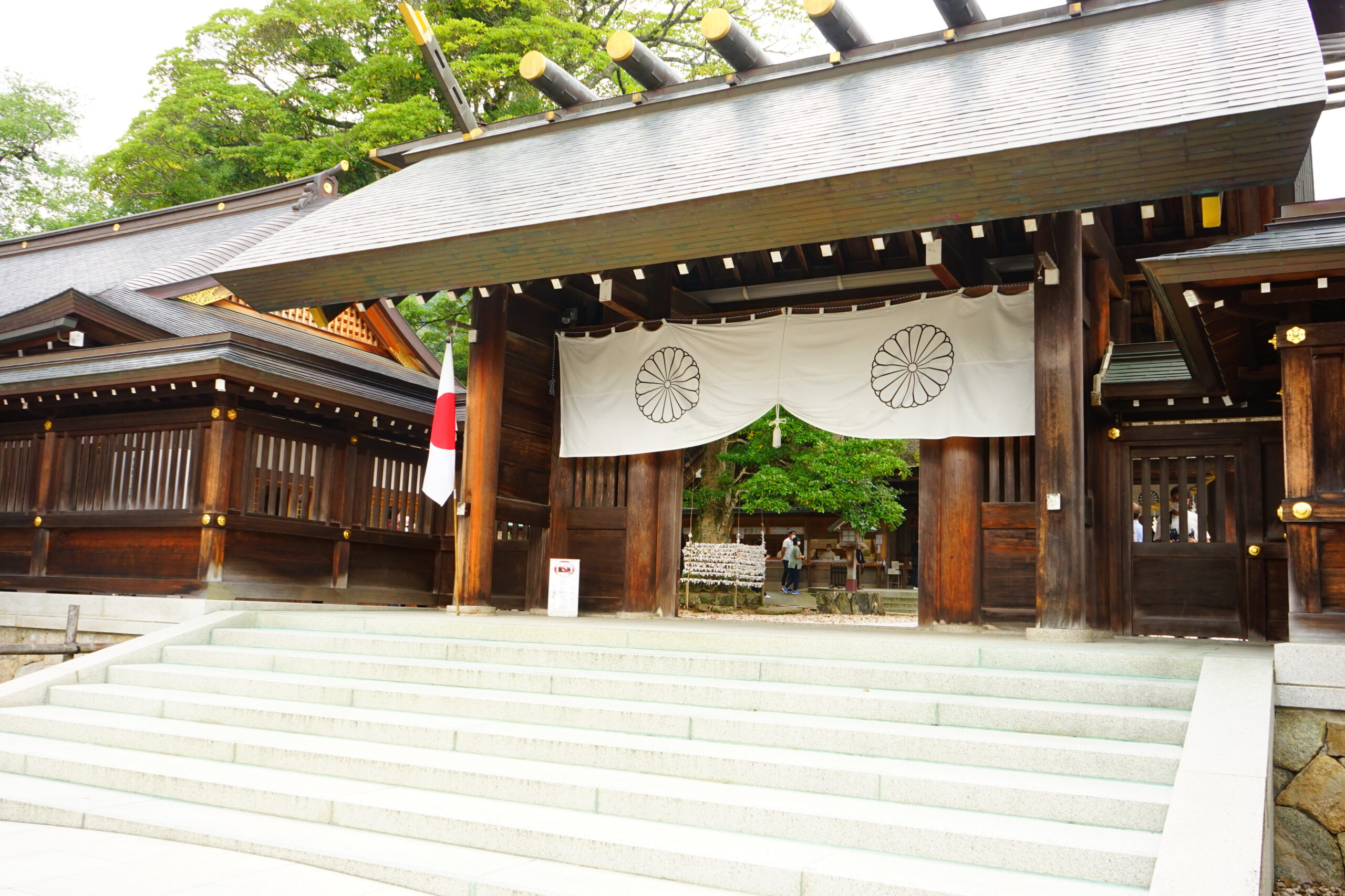 Front view of a traditional Japanese Shinto shrine entrance with dark wooden beams, white banners featuring chrysanthemum crests, and a nearby Japanese flag beside the steps amid green trees.