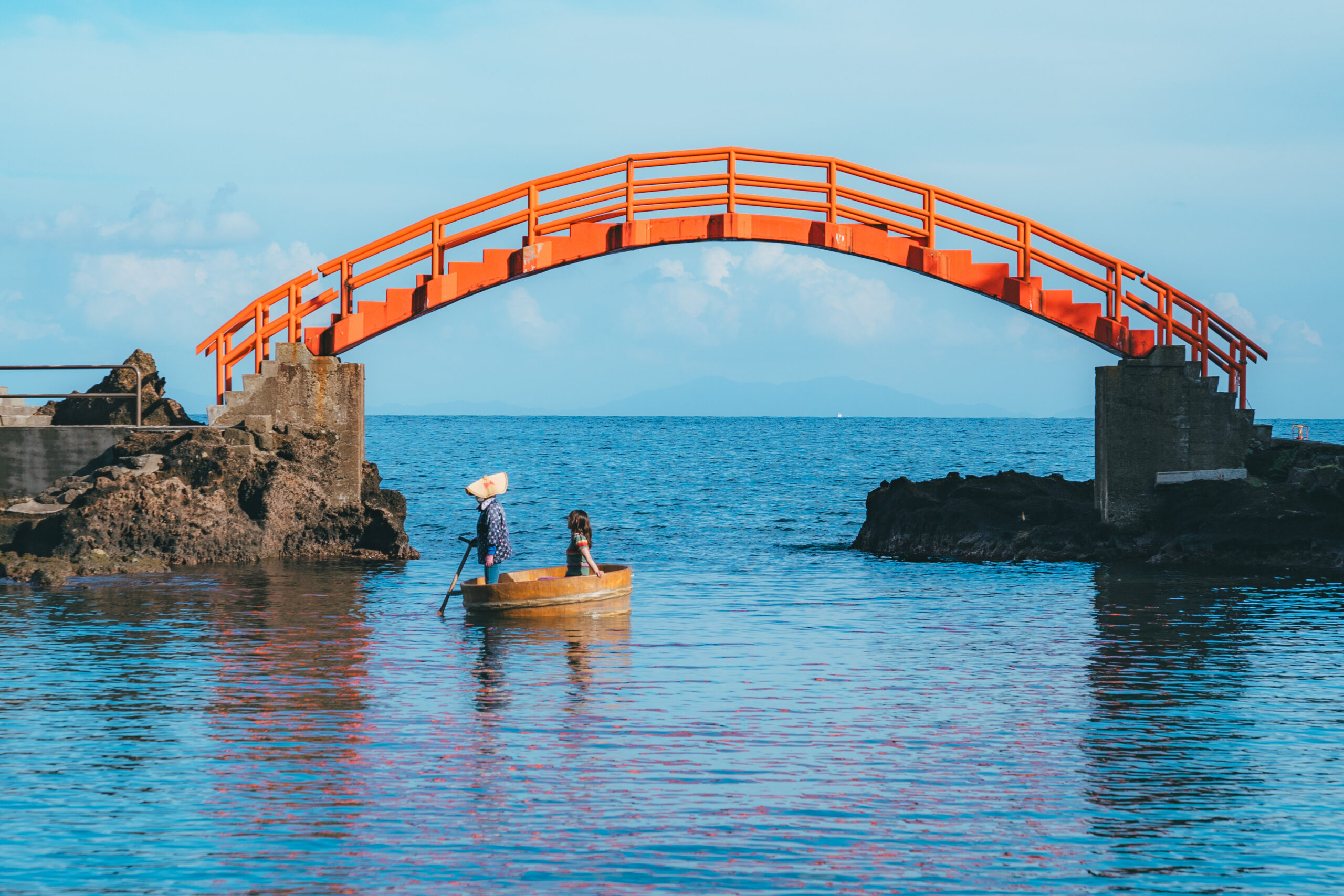 Bright orange curved footbridge spans the blue sea, with two people in a small boat near the rocky shore below.