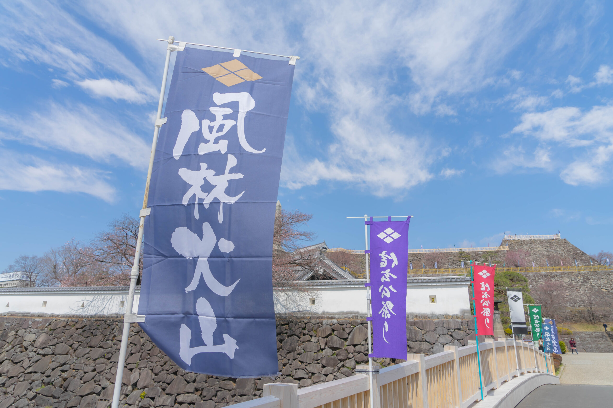 Blue vertical banner with white Japanese characters fluttering beside a stone wall; other purple, red, and green banners line a pedestrian walkway under a clear blue sky.