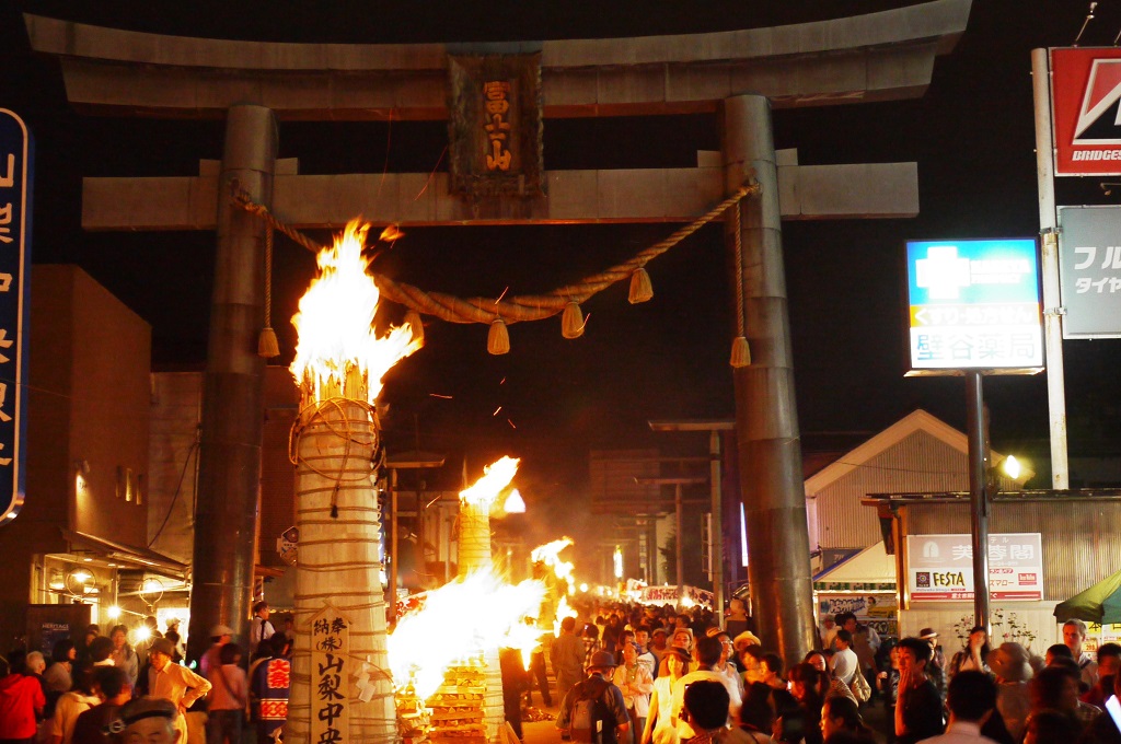 Nighttime festival at a Shinto torii gate with tall blazing torches and a crowded street behind.