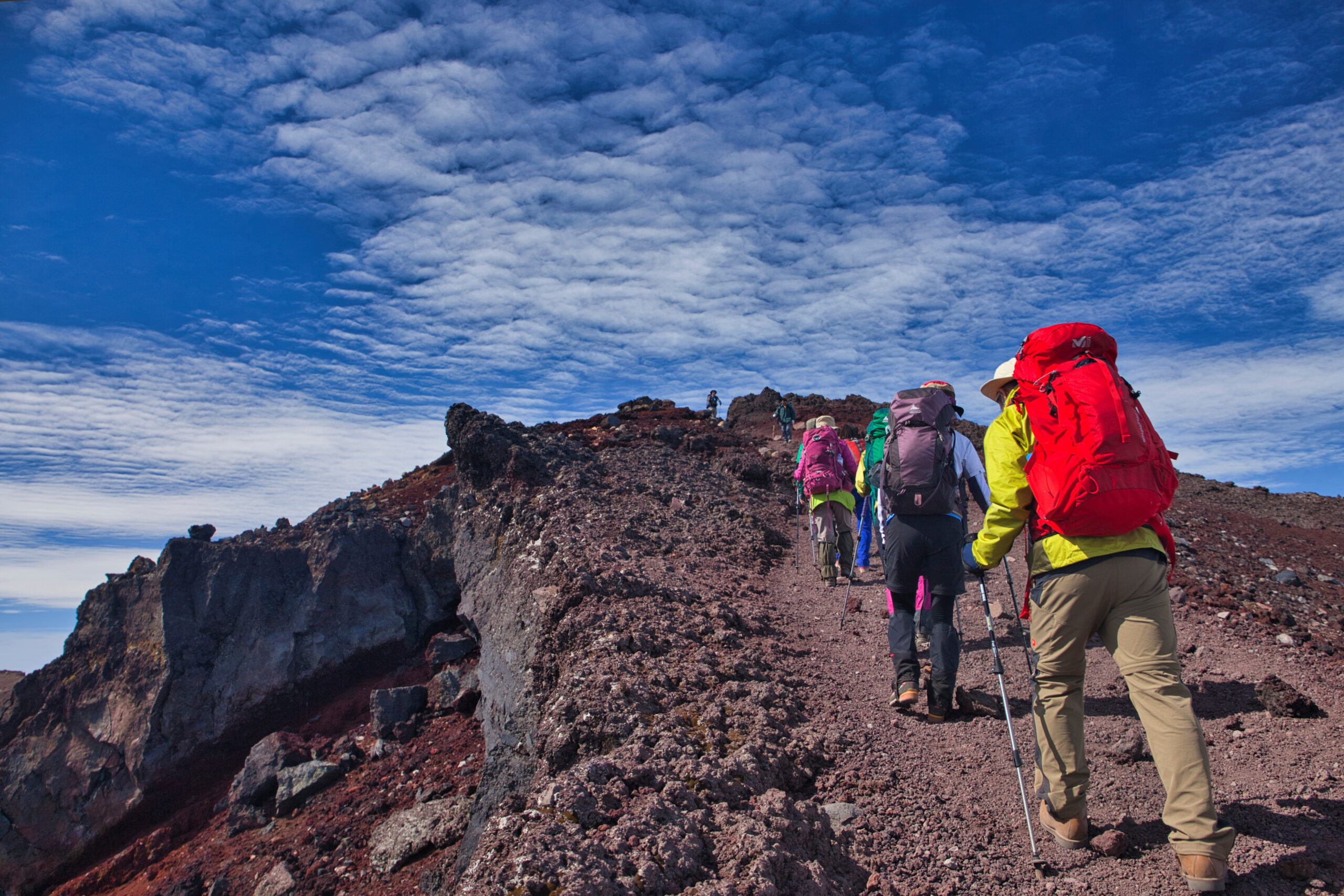 Group of hikers with colorful backpacks and trekking poles ascending a rocky mountain trail under a bright blue sky, approaching a summit.