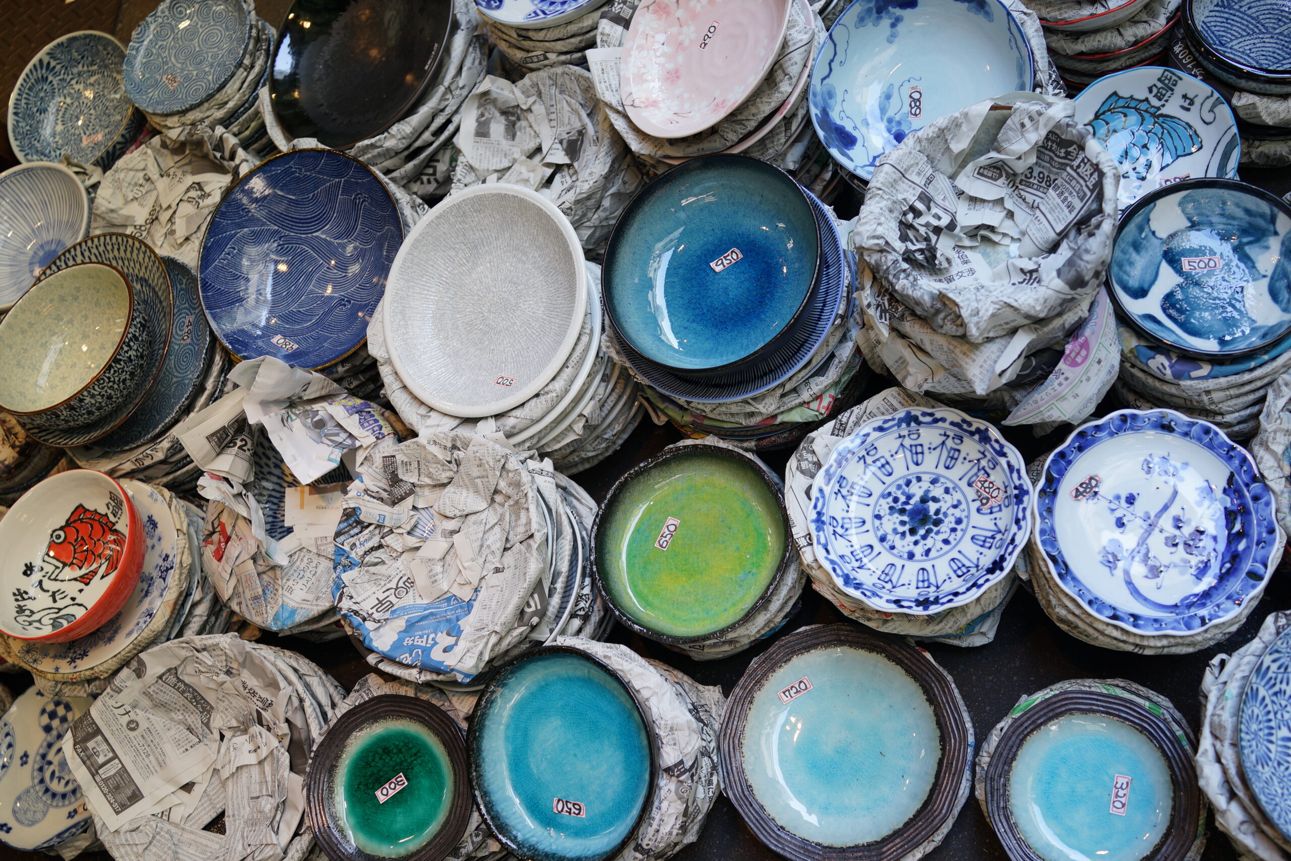 Pile of ceramic bowls and plates with price stickers, nestled in crumpled newspaper in a market display.