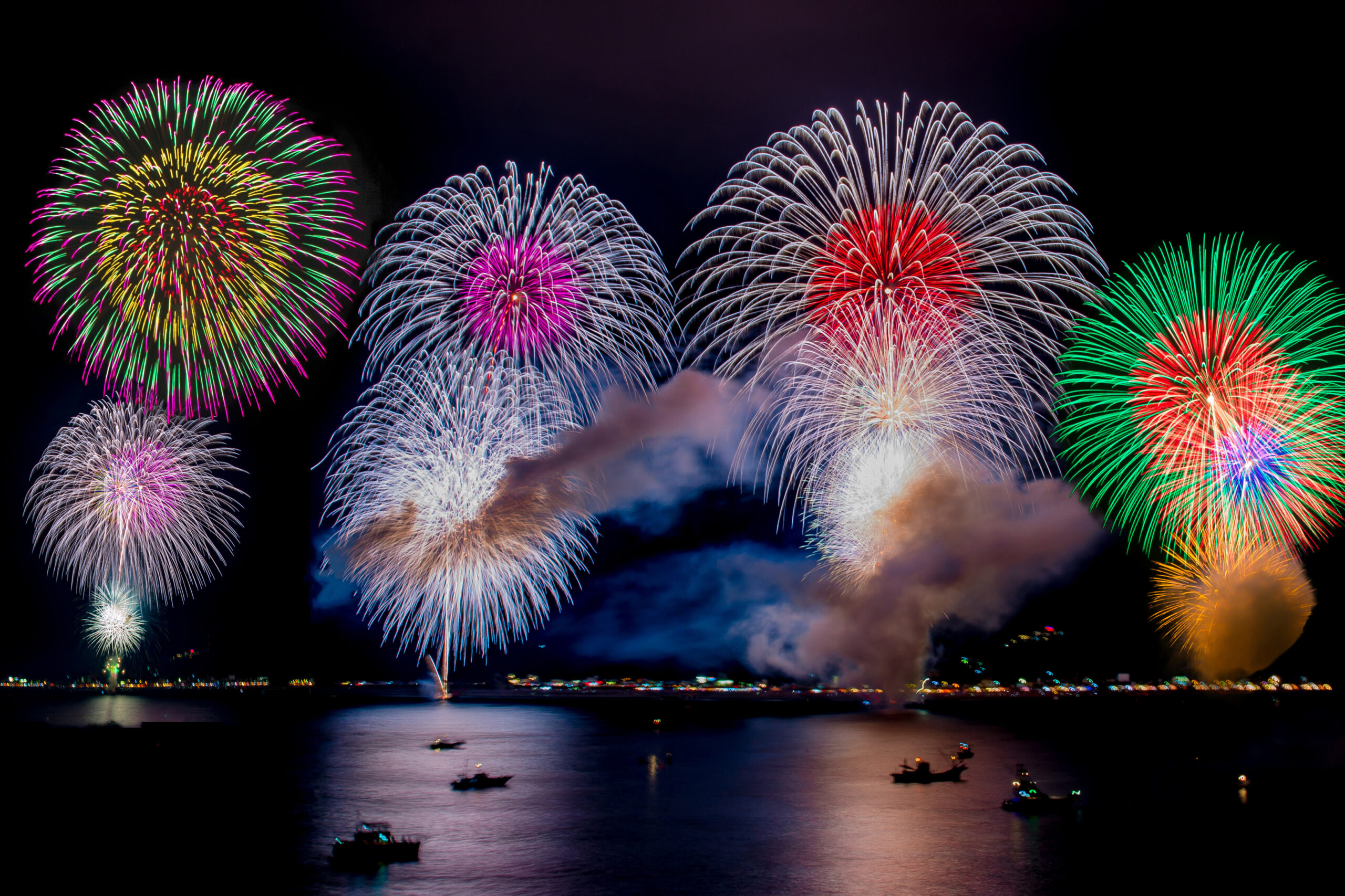 Colorful fireworks bursting over a dark night ocean, with reflections on the water and small boats on the shore.