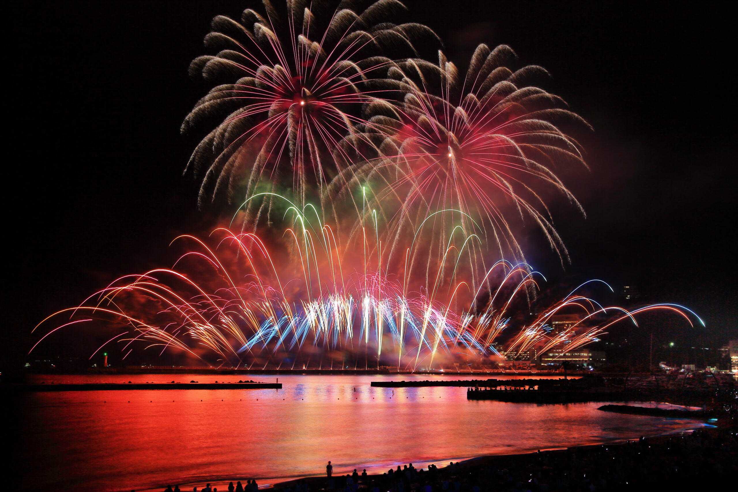 Fireworks exploding in pink and red above a dark harbor, reflected in the water with silhouettes watching from the shore.