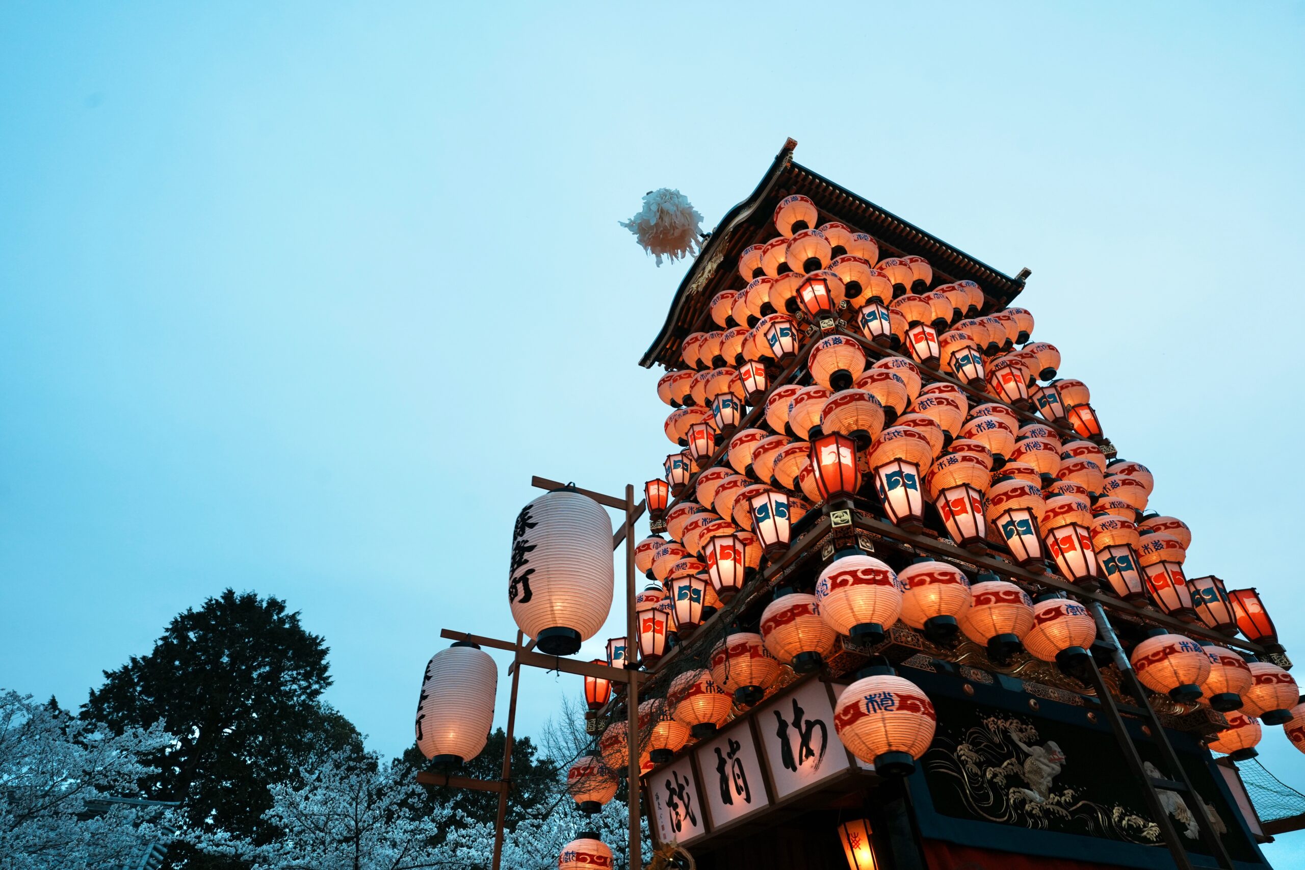 Towering festival structure adorned with hundreds of red and white lanterns against a clear blue sky, with a pagoda-style roof at the top and illuminated signs in kanji/Chinese characters.