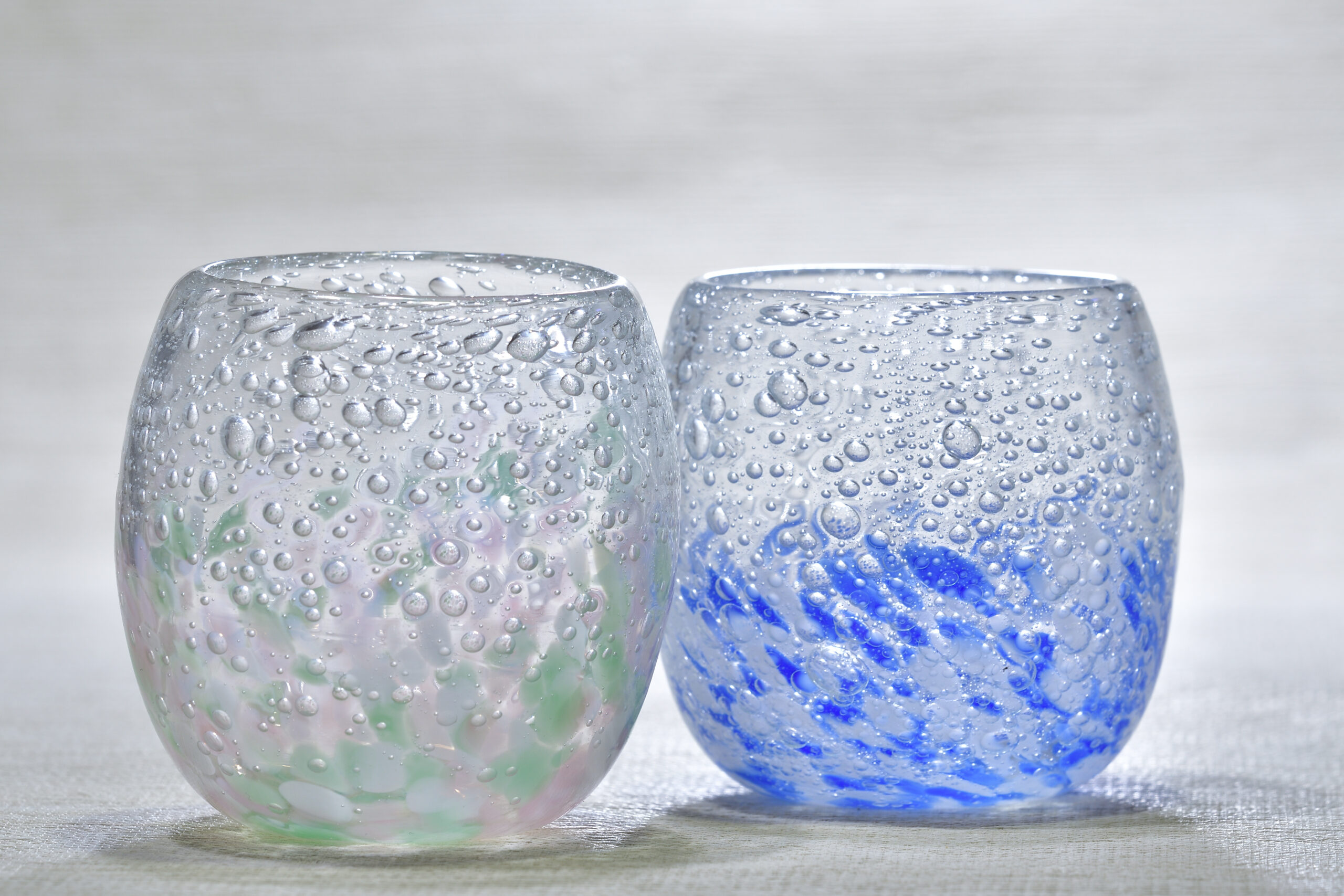 Two glass tumblers with condensation droplets, one with a green-tinted base and the other blue, resting on a light surface.