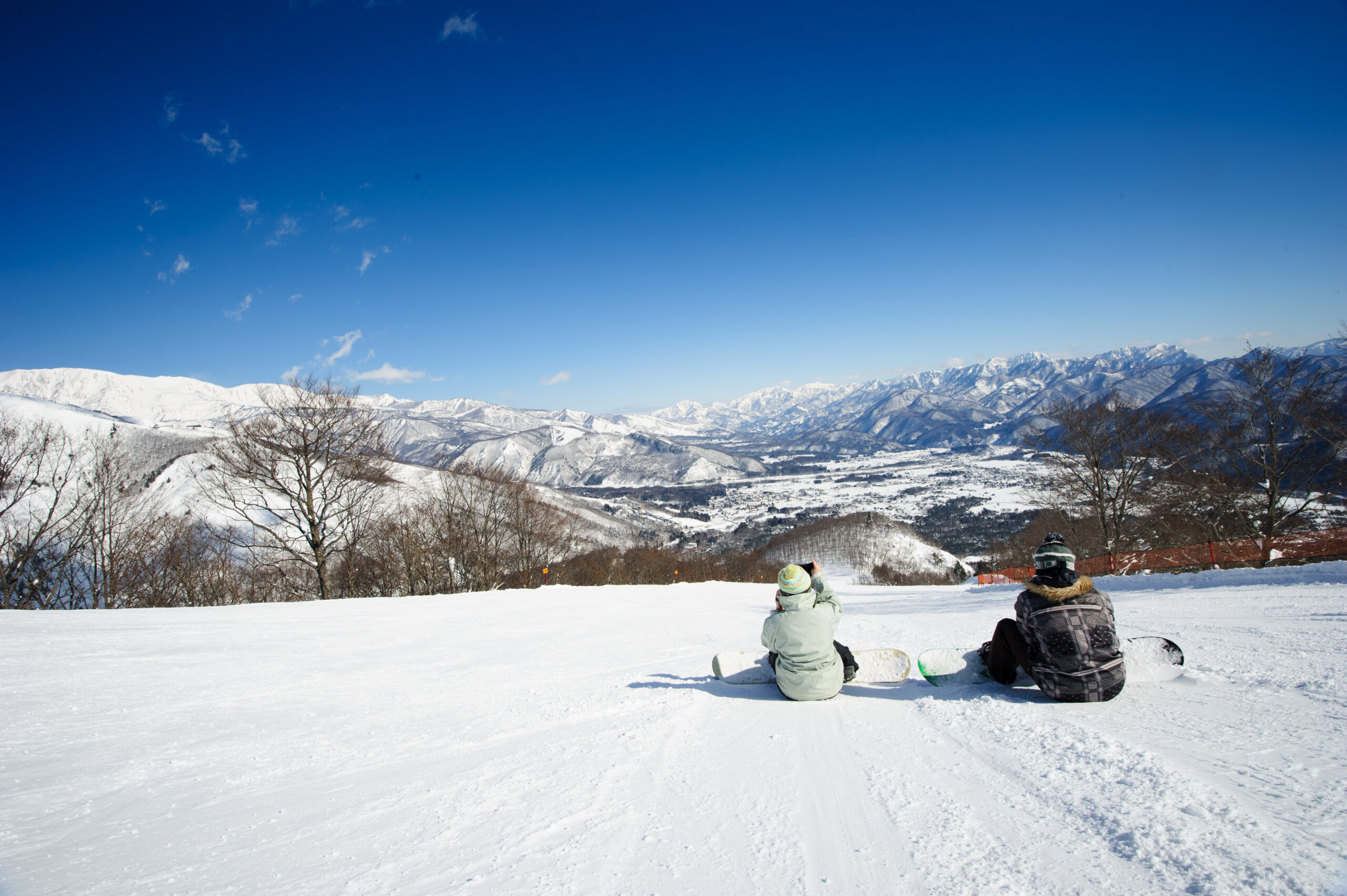 Snowy mountain landscape with two people on snowboards resting in the foreground under a clear blue sky.