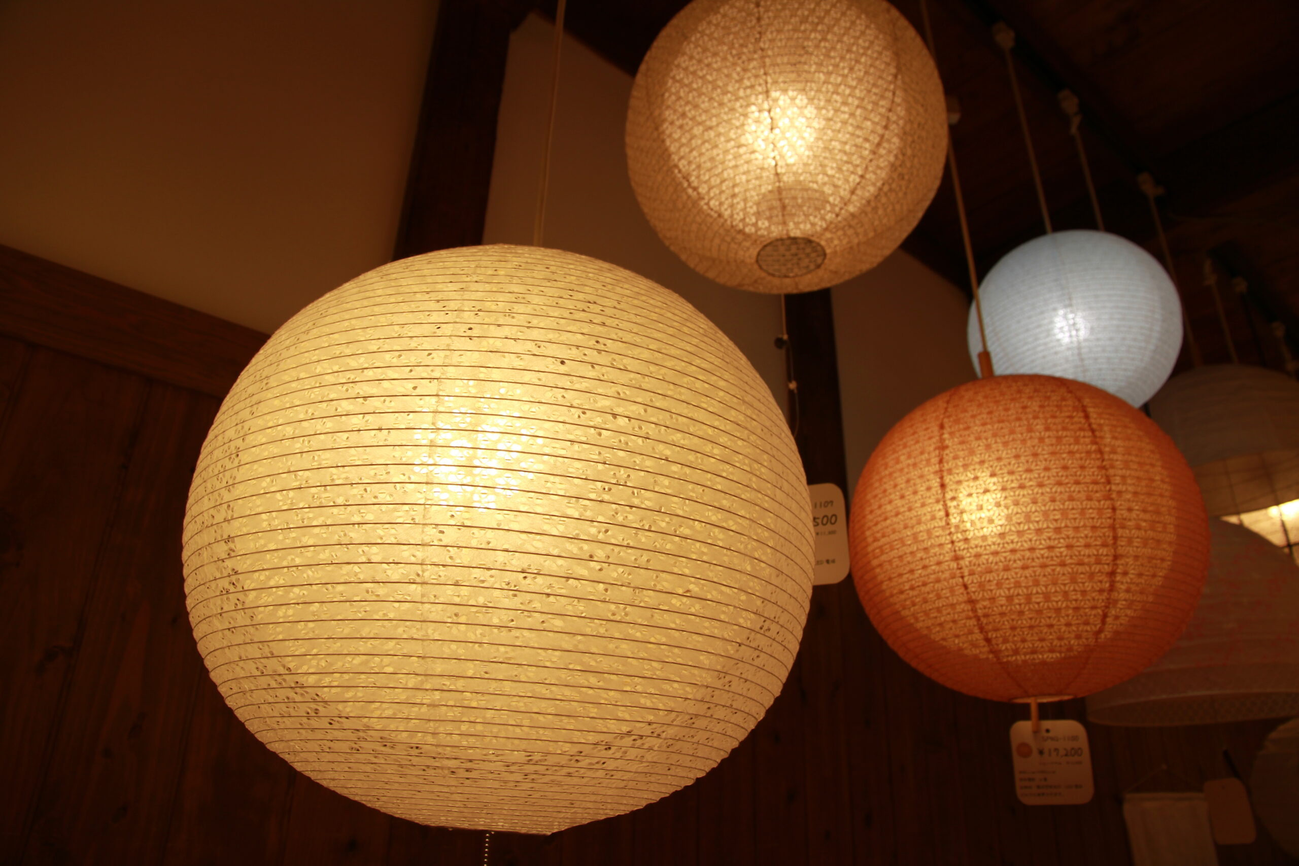 Warm-lit paper lanterns in cream, orange, and blue hanging from cords against a wooden wall in a shop setting.