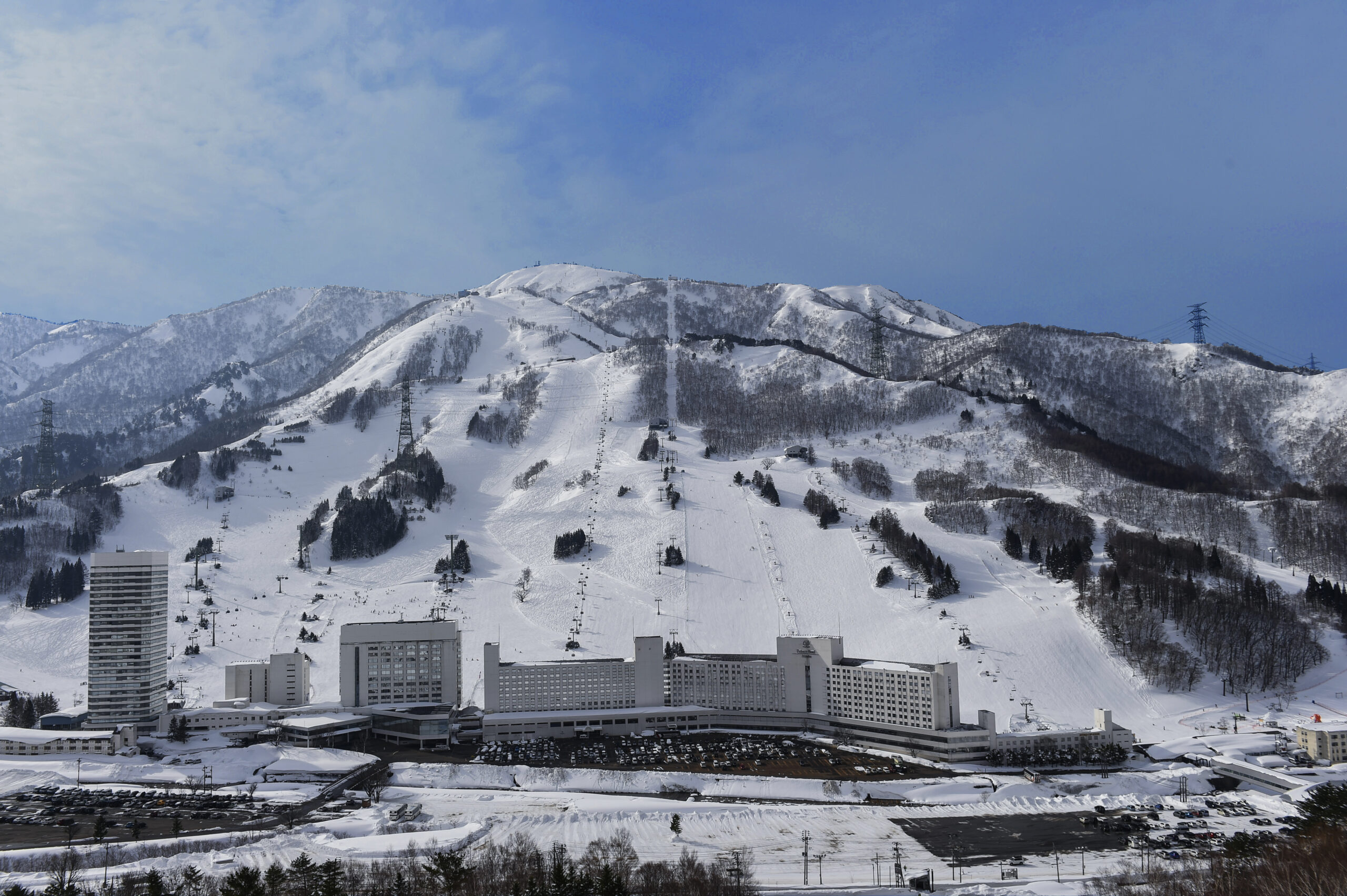 Snow-covered mountains loom behind a cluster of modern buildings at the base, with ski slopes and chairlifts visible on the hillside.