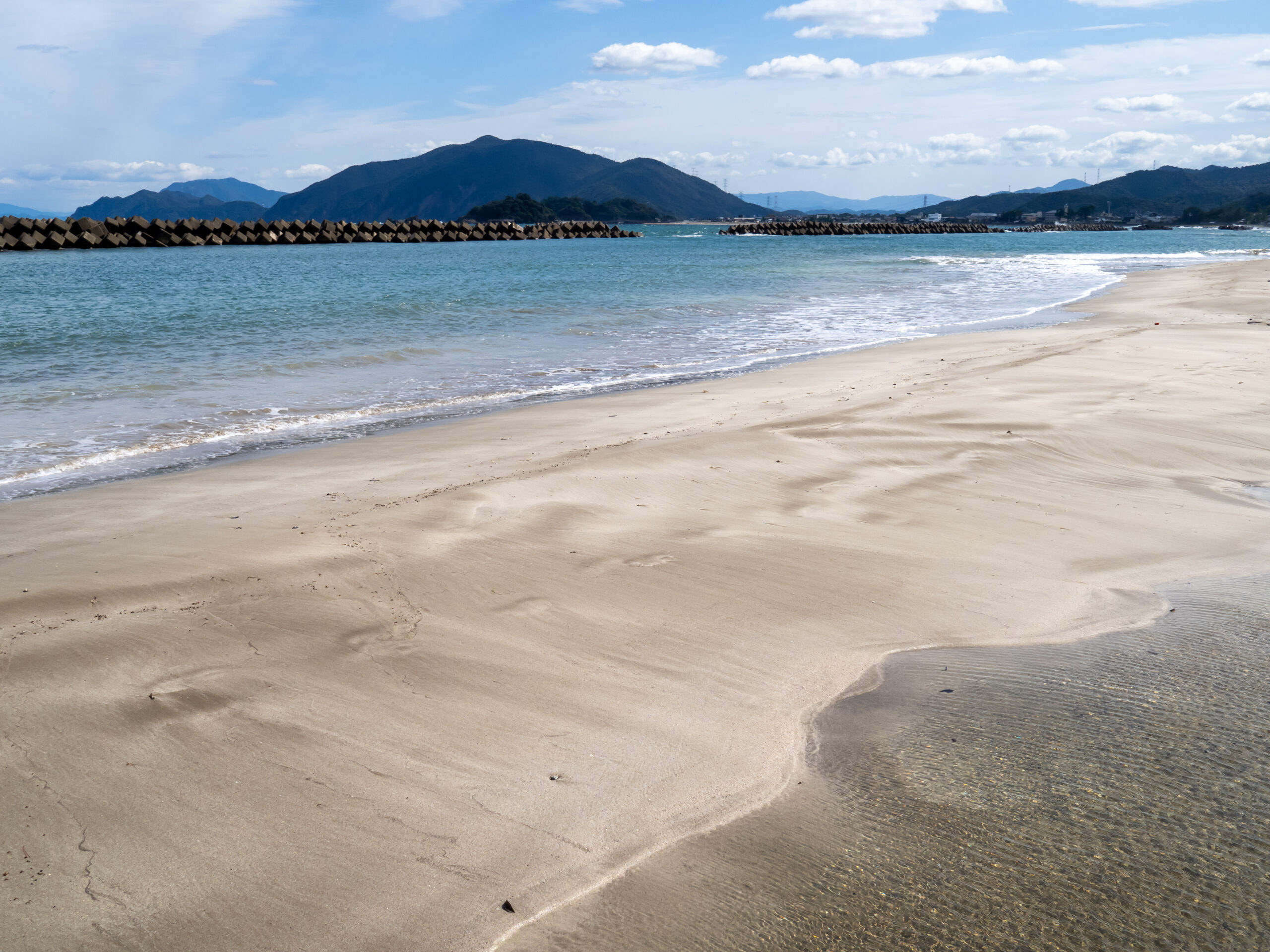 Quiet beach with smooth wet sand, gentle waves, and a breakwater and mountains on the horizon under a blue sky as clouds drift by.