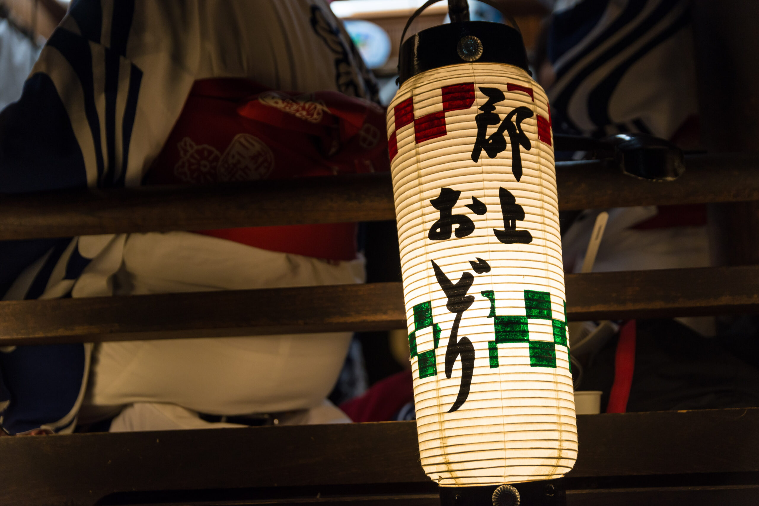 Japanese paper lantern with black kanji characters, hanging between wooden beams, warm festival lighting behind it.
