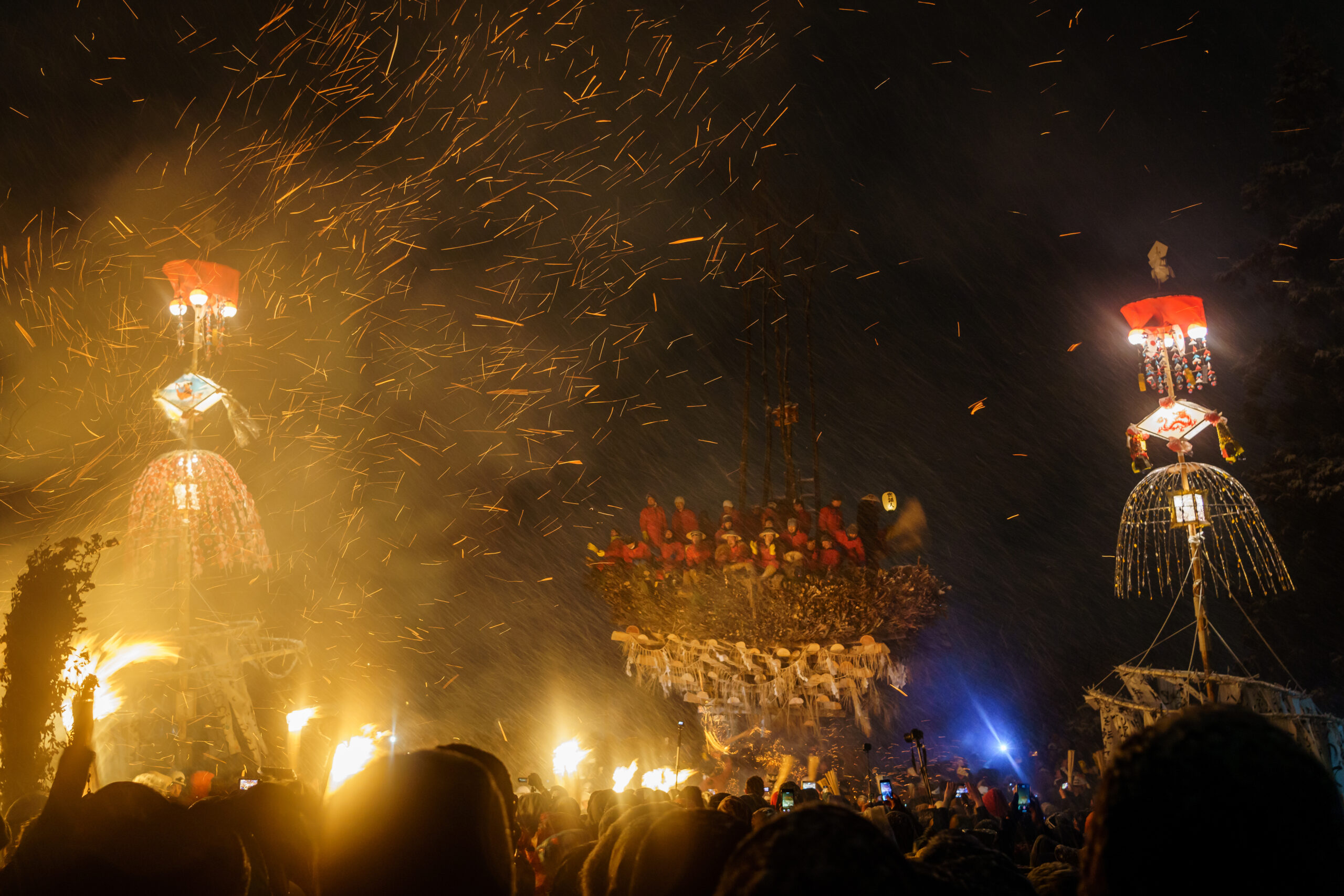 Night festival scene with glowing lanterns, fire columns, and sparks flying in the dark as a crowd watches from below.
