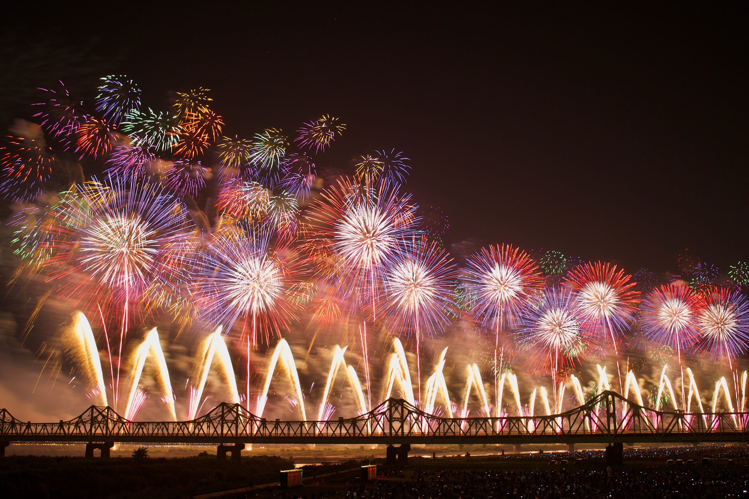 Colorful fireworks burst over a dark night sky above a silhouetted bridge, while a crowd watches from the riverbank.