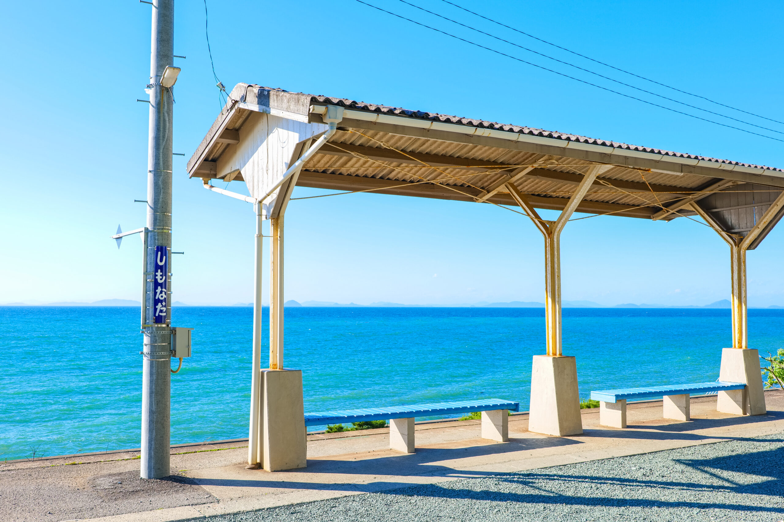 Open-air seaside shelter with metal columns overlooking the bright blue ocean and distant horizon under a clear sky on a sunny day.