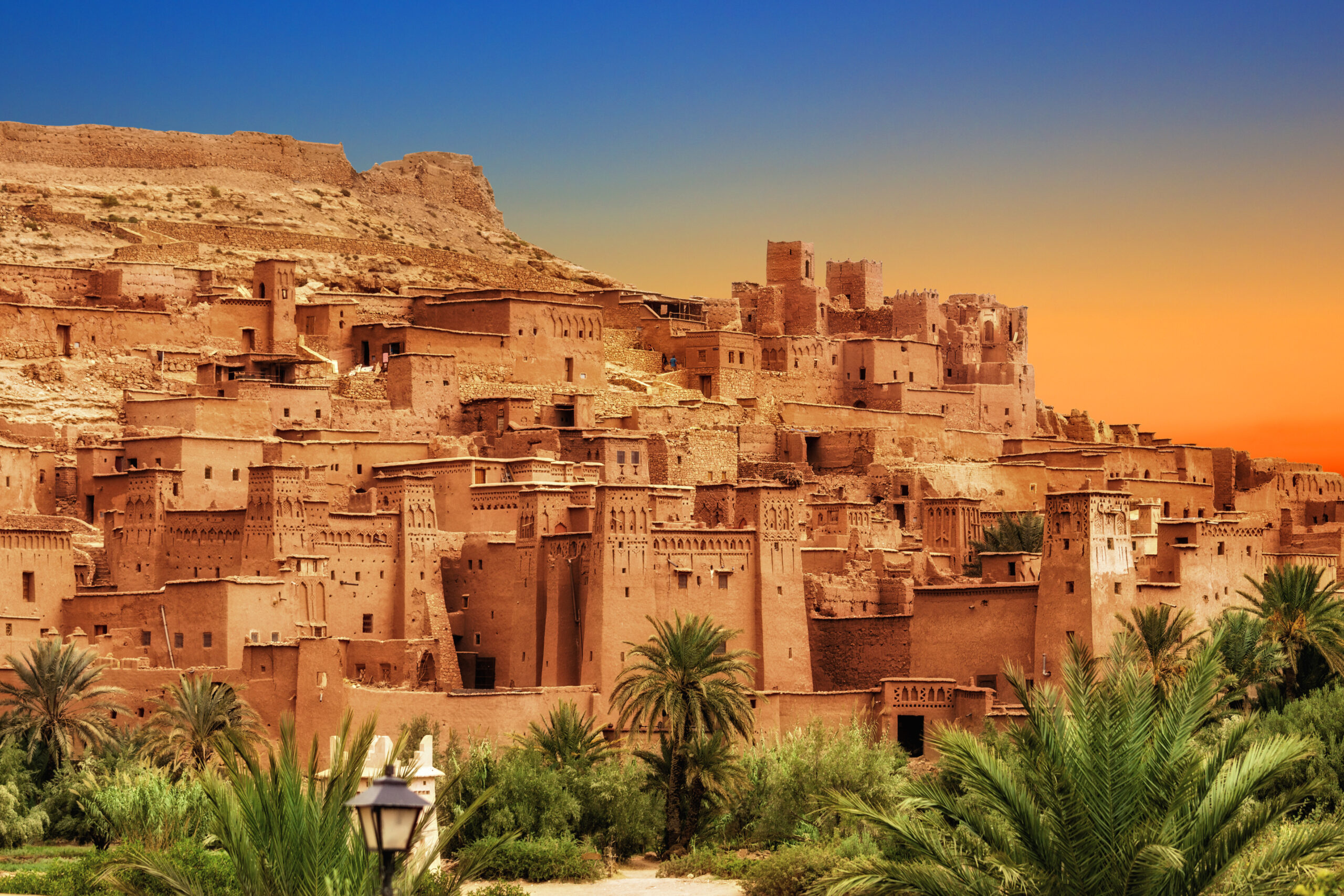 Desert kasbah town of clay buildings on a hill at sunset, with palm trees in the foreground.