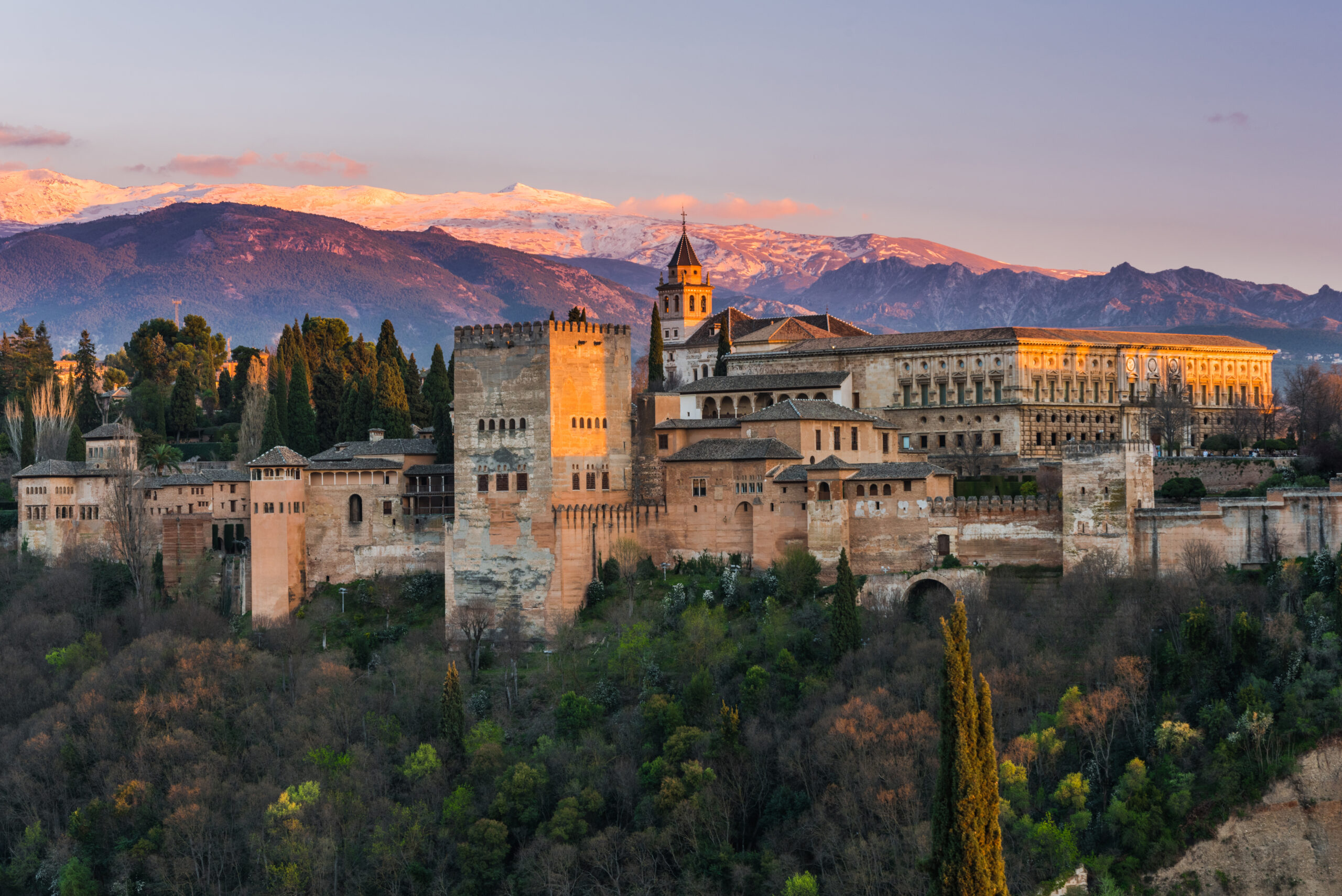 The Alhambra fortress and palace complex atop a hill, with a forested foreground and snow-capped Sierra Nevada mountains in the distance at sunset.