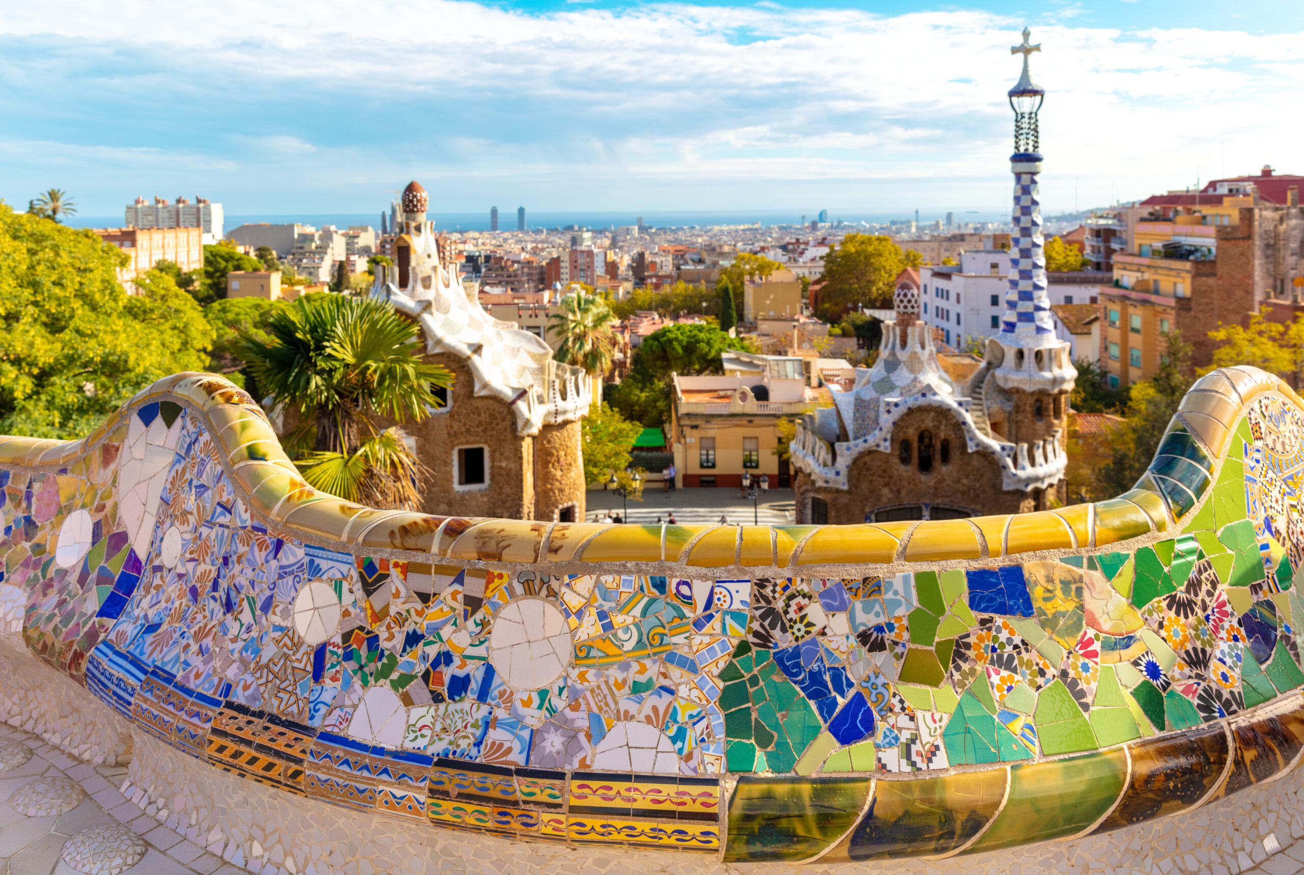 Colorful mosaic serpentine bench at Park Güell with Barcelona skyline and sea in the distance.