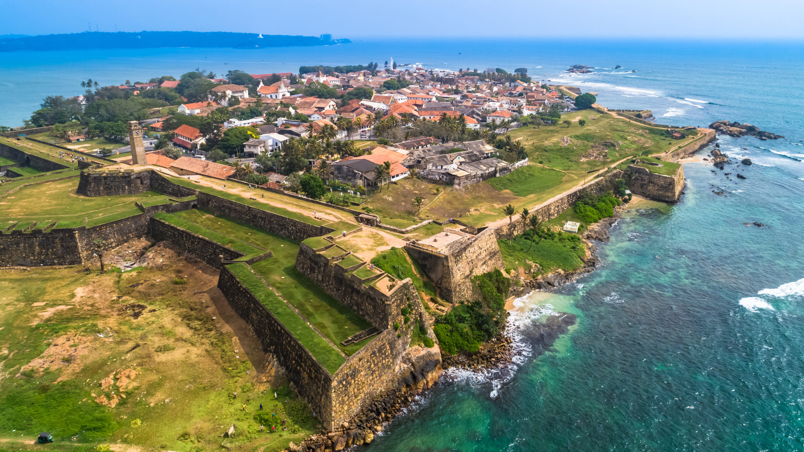 Aerial view of a coastal town with red-roofed houses inside historic fortifications along a rocky shoreline and turquoise sea.