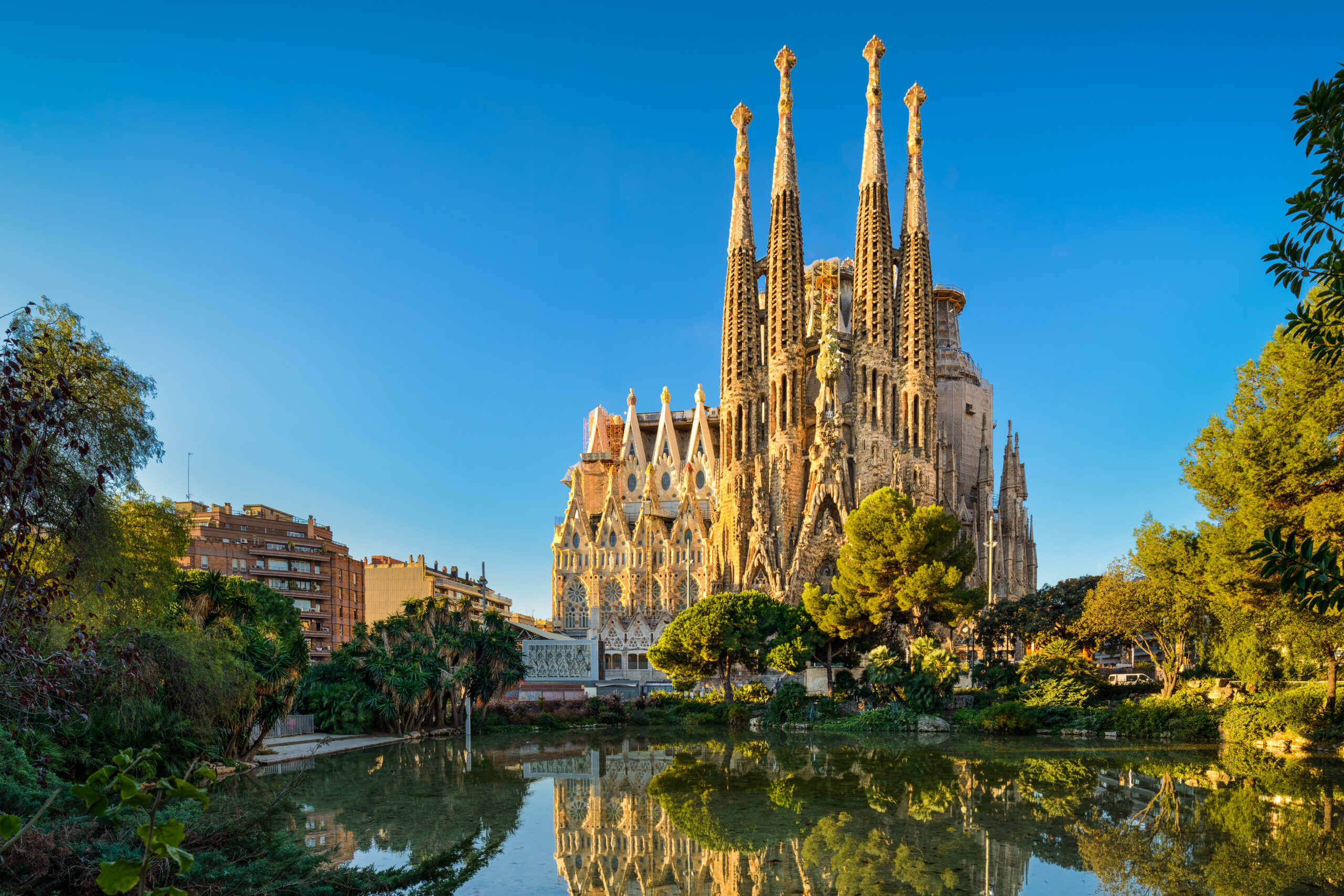 Sagrada Família's tall spires rise beside trees under a clear blue sky, mirrored in a calm river.]