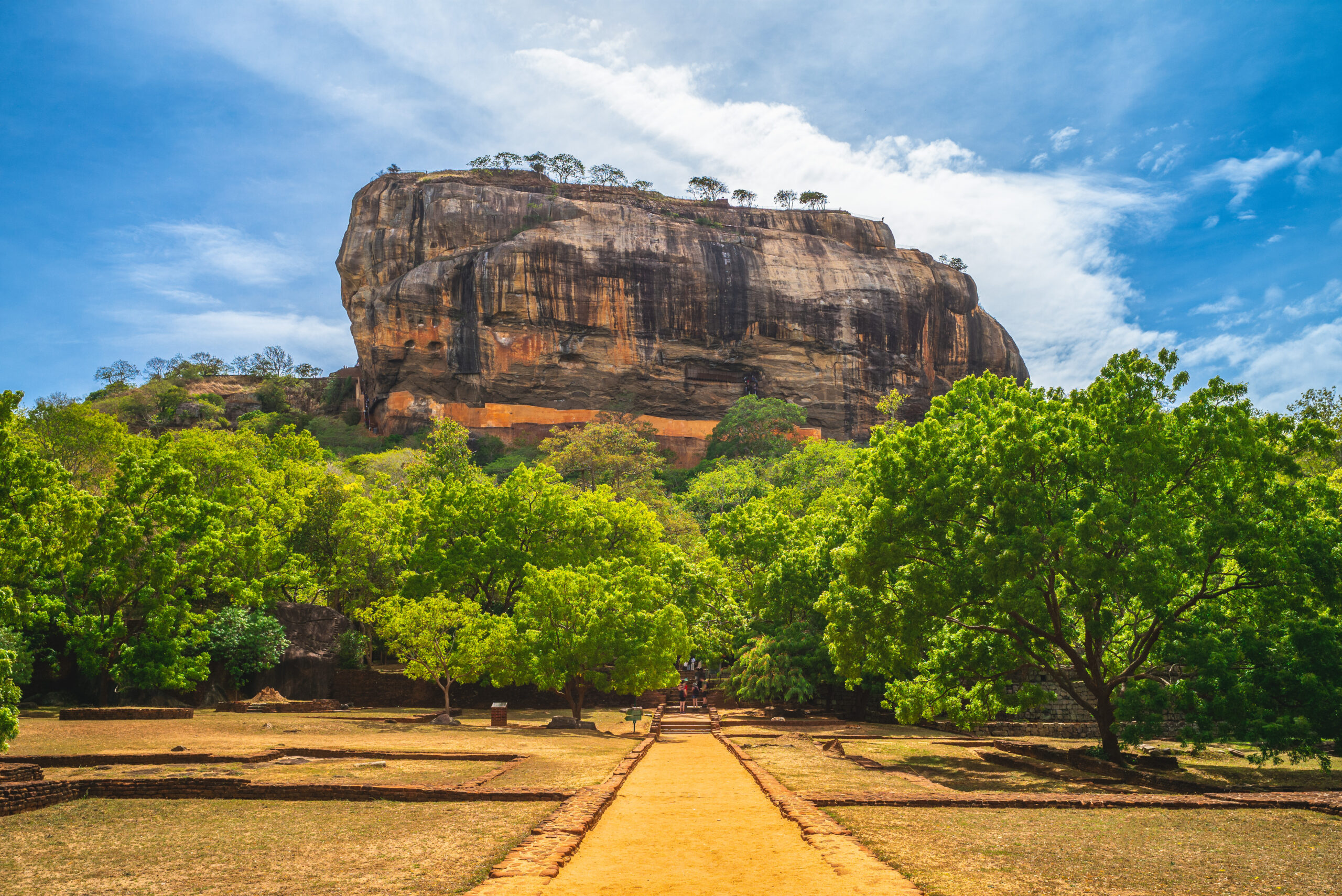 Massive rock cliff topped with trees, with a sandy path and stone edging in the foreground.